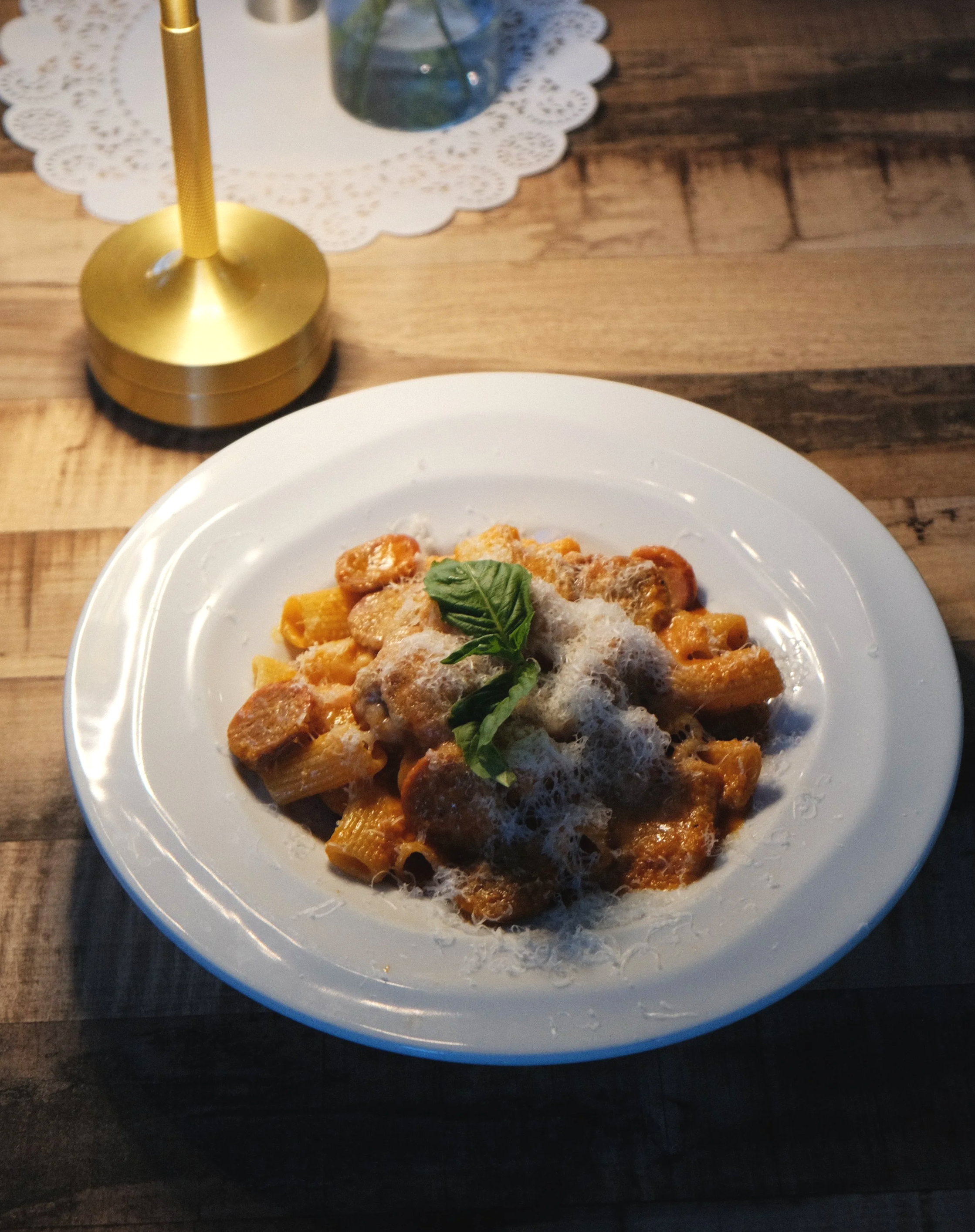 A plate of pasta with sauce, topped with grated cheese and basil, on a wooden table. In the background, there is a gold-colored lamp and a glass of water on a doily.