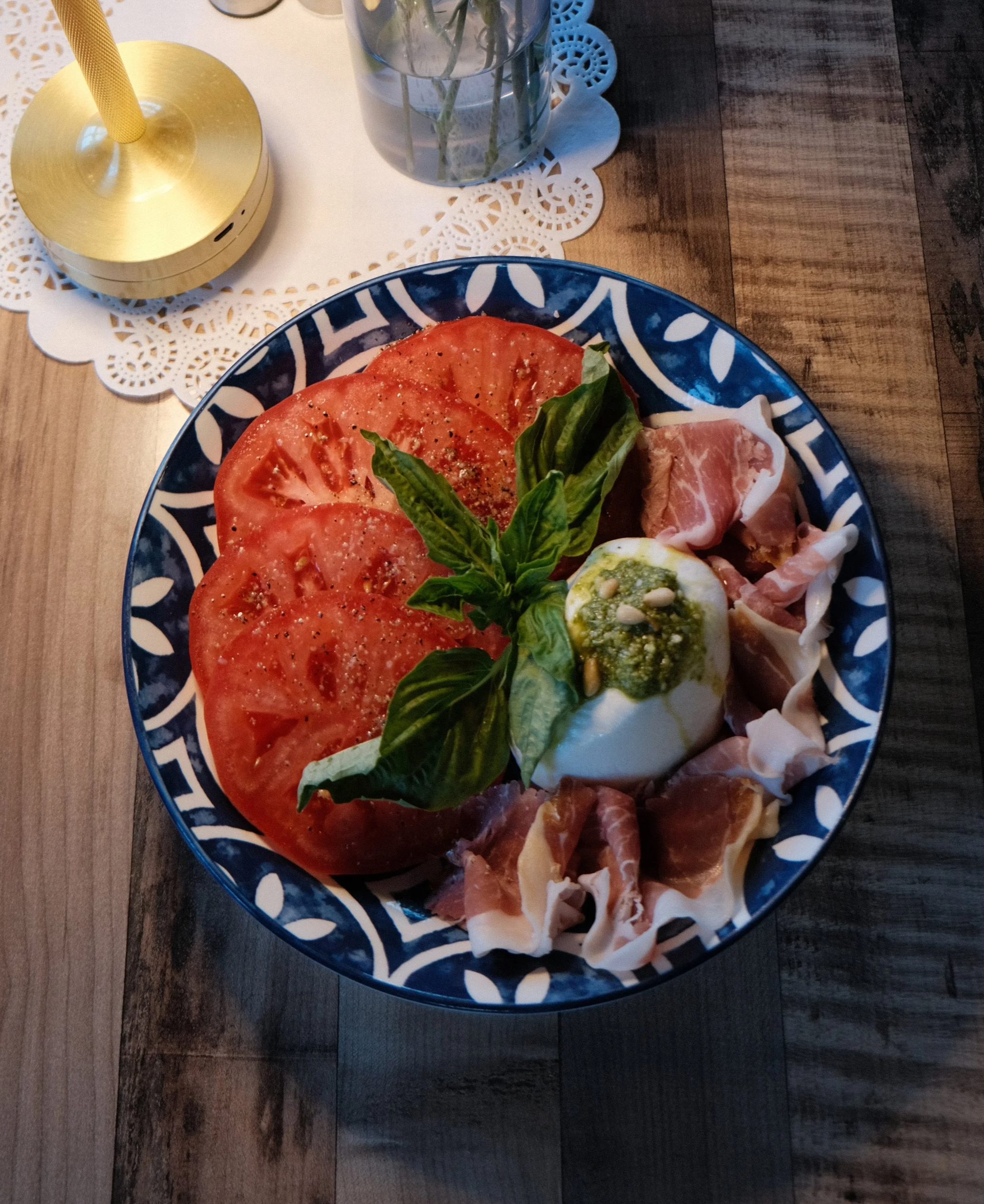 A plate of burrata cheese with sliced tomatoes, fresh basil leaves, and prosciutto on a decorative blue and white plate, placed on a wooden table. In the background, there is a glass of water with stems of a plant and a gold-colored table lamp.