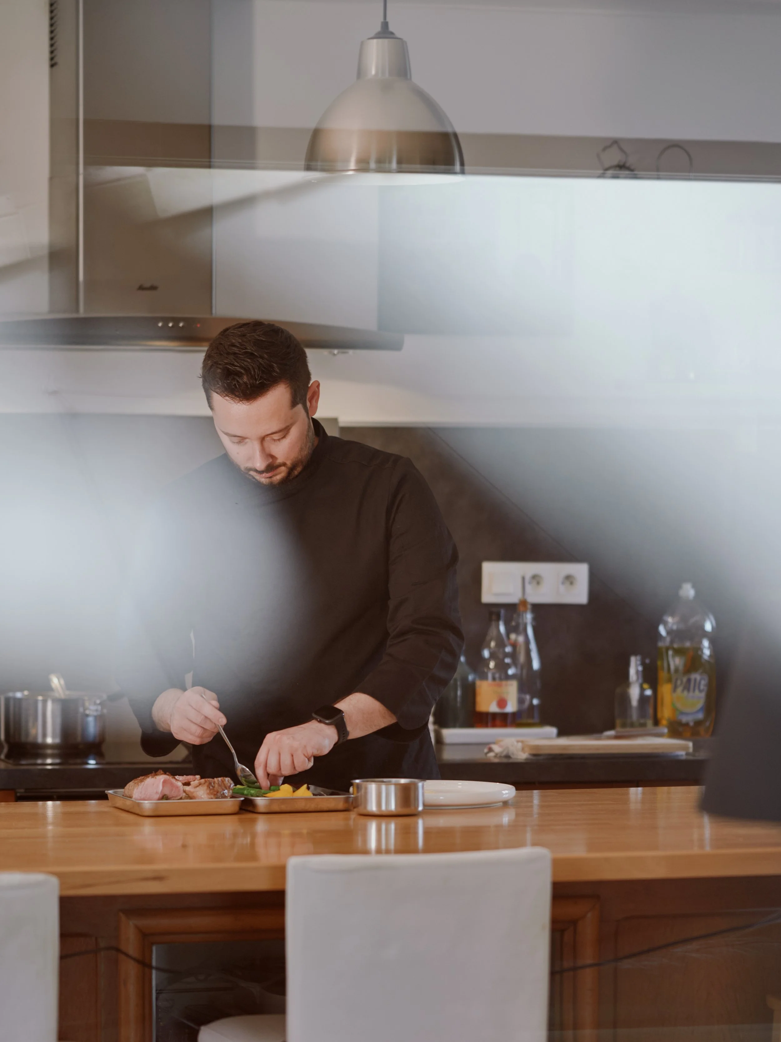 Un chef prépare un repas dans une cuisine moderne, avec des bouteilles et des ingrédients sur le plan de travail en bois.