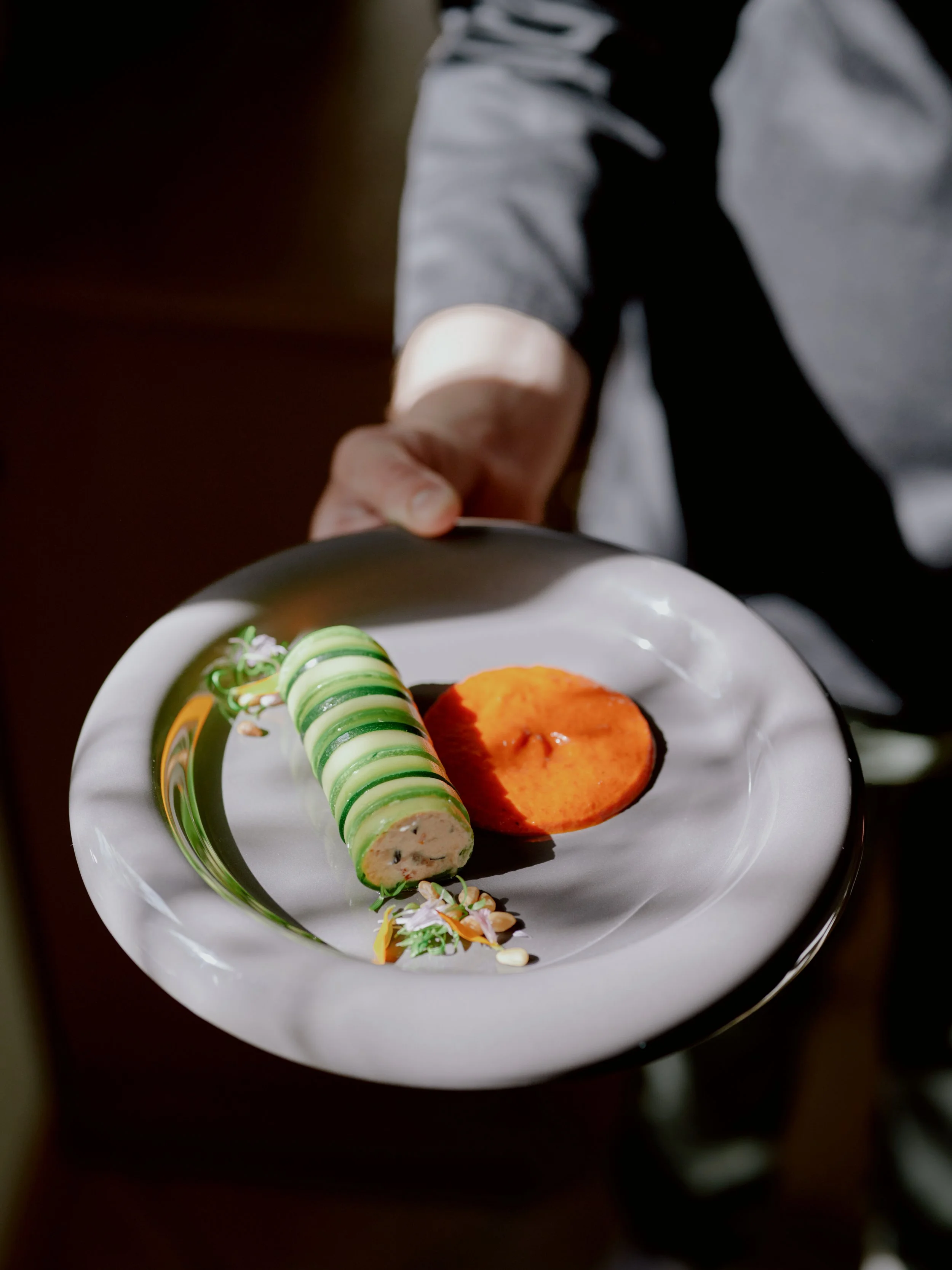 Un plat de nourriture présenté sur une assiette blanche, comprenant un rouleau de courgette avec de la brousse à  l'intérieur et une sauce aux poivrons.