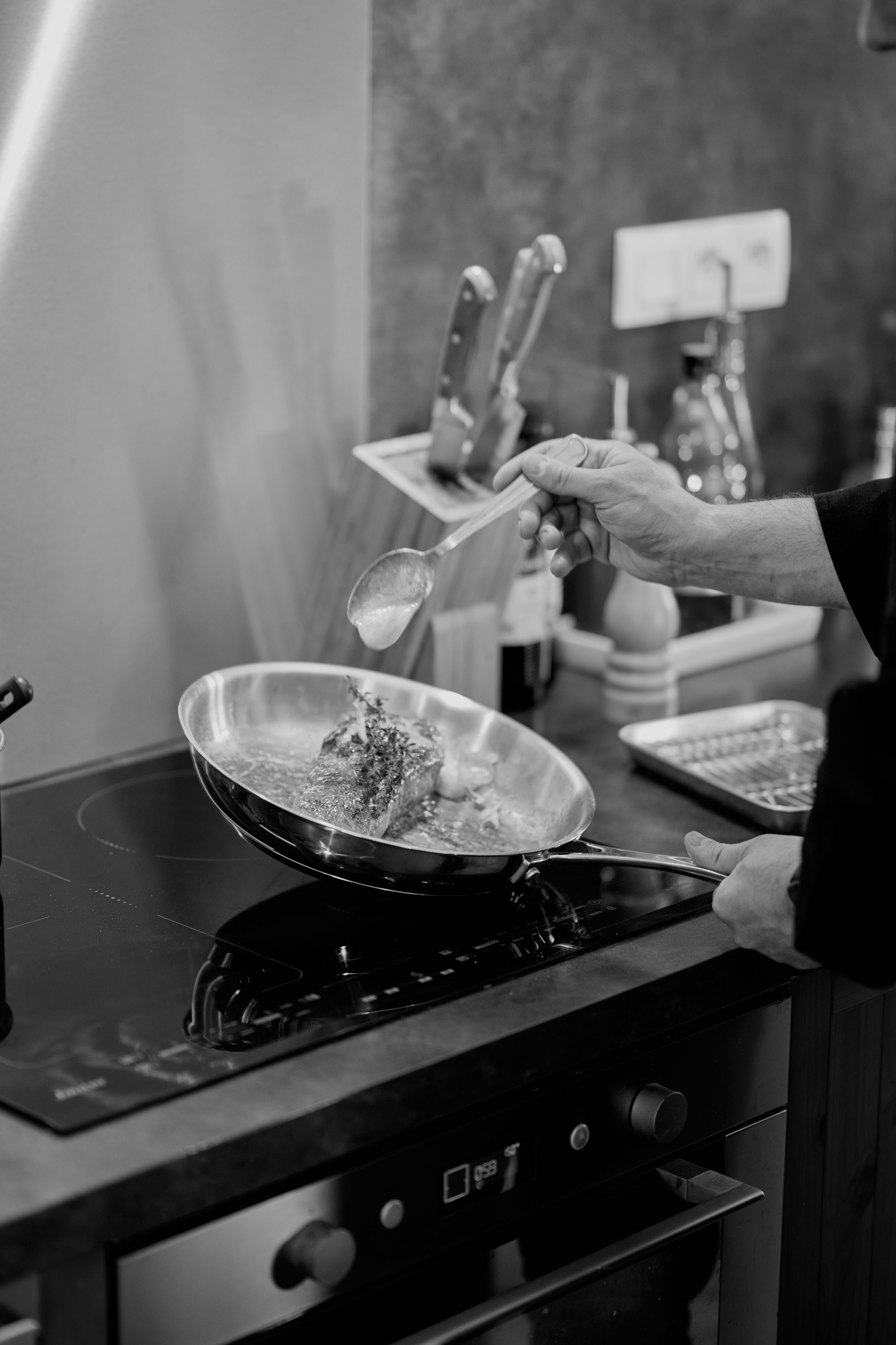 Une personne en train de cuisiner, versant une sauce ou un liquide dans une poêle sur une cuisinière électrique. Il y a des ustensiles suspendus en arrière-plan, une bouteille d'eau et un évier.