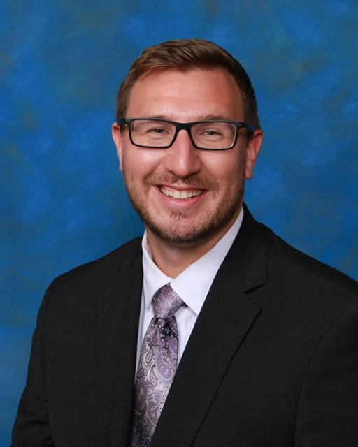 Portrait of a smiling man with glasses, wearing a black suit, white shirt, and patterned tie, against a blue background.