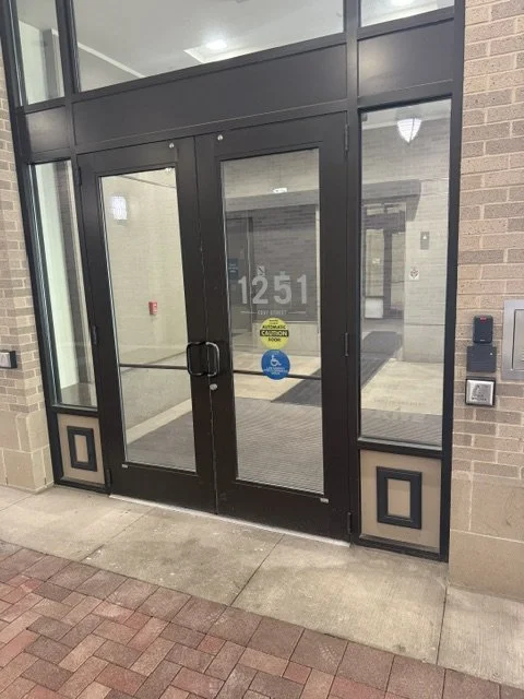 Glass double doors with black frames, door number 1251, and accessibility symbols, at the entrance of a building.