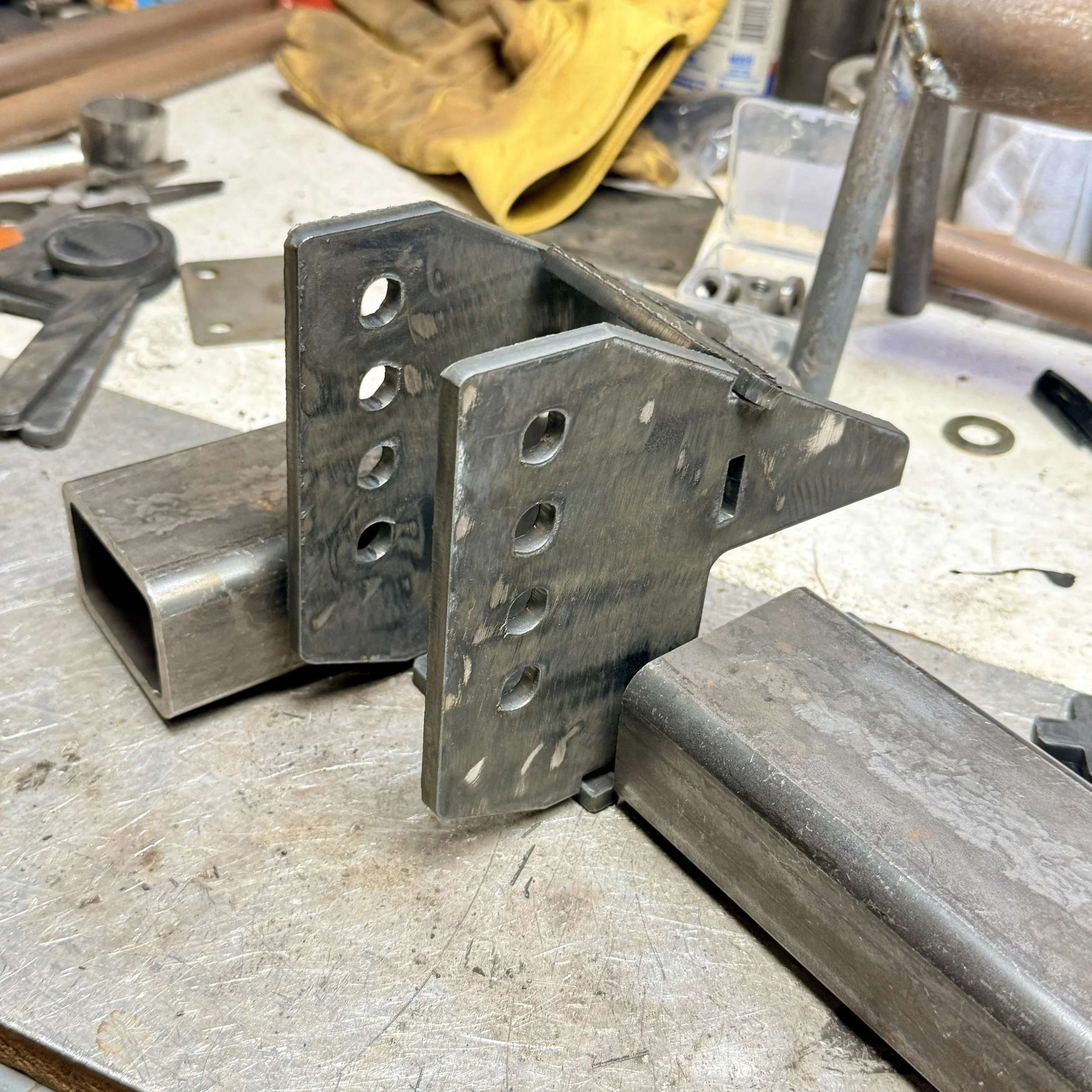 Close-up of metal workpieces and tools on a workbench, with some yellow gloves in the background.