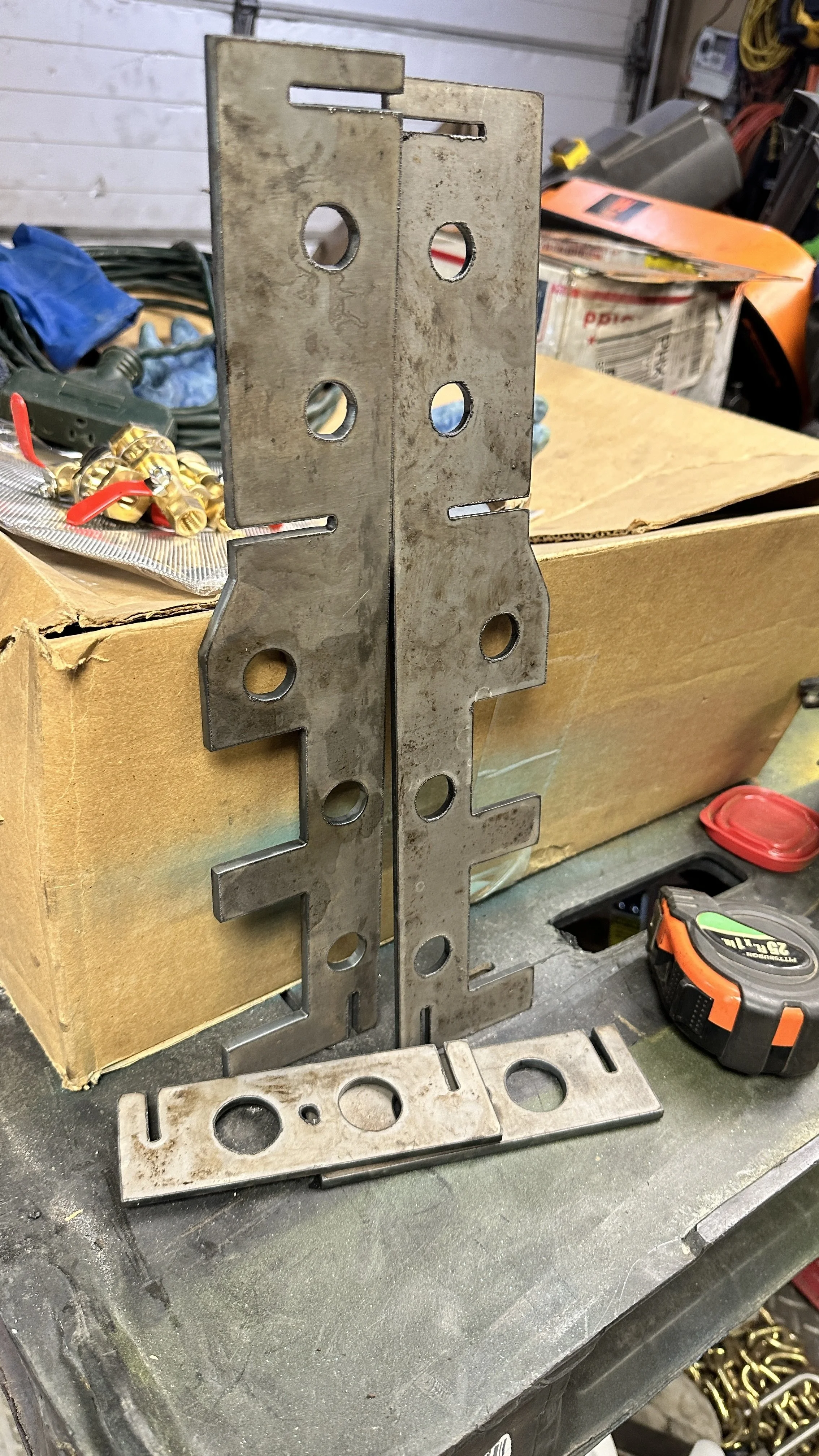 Metal bracket with multiple holes and cutouts, standing upright on a workbench in a workshop.