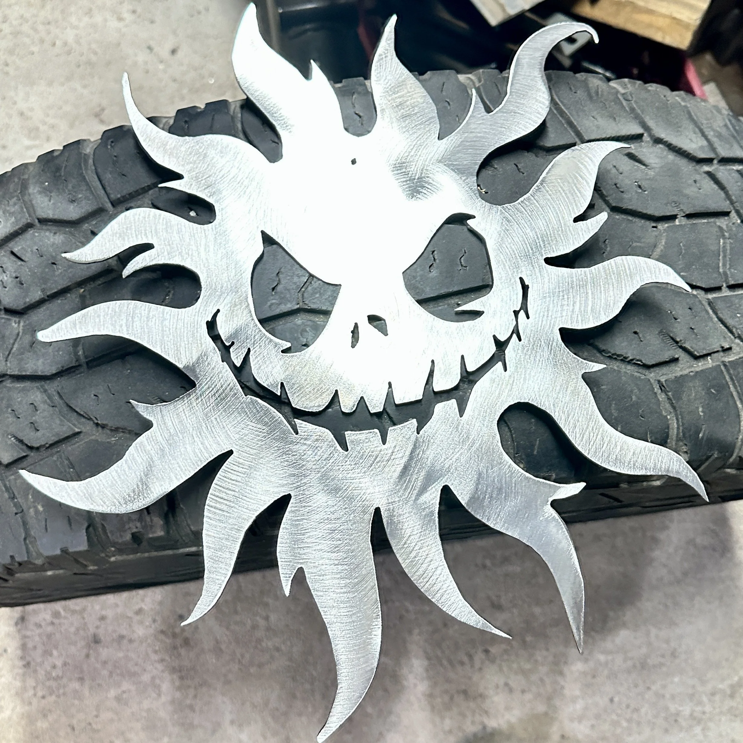 A silver metal skull emblem placed on a tire with a textured surface. The emblem has sharp, jagged features resembling a stylized skull.