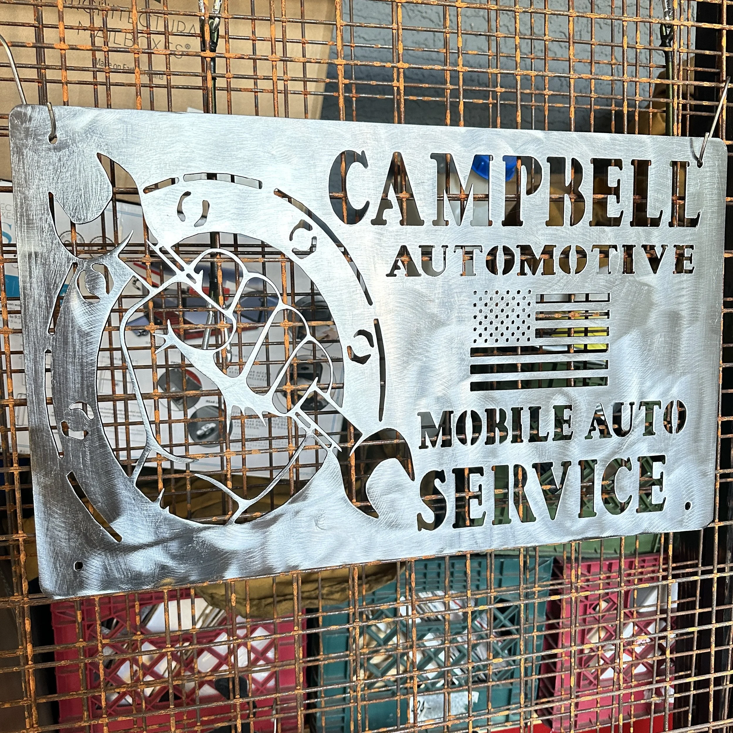A metal sign with cut-out text and a bicycle illustration hanging on a rusted wire grid rack, advertising an automotive and self-service shop.
