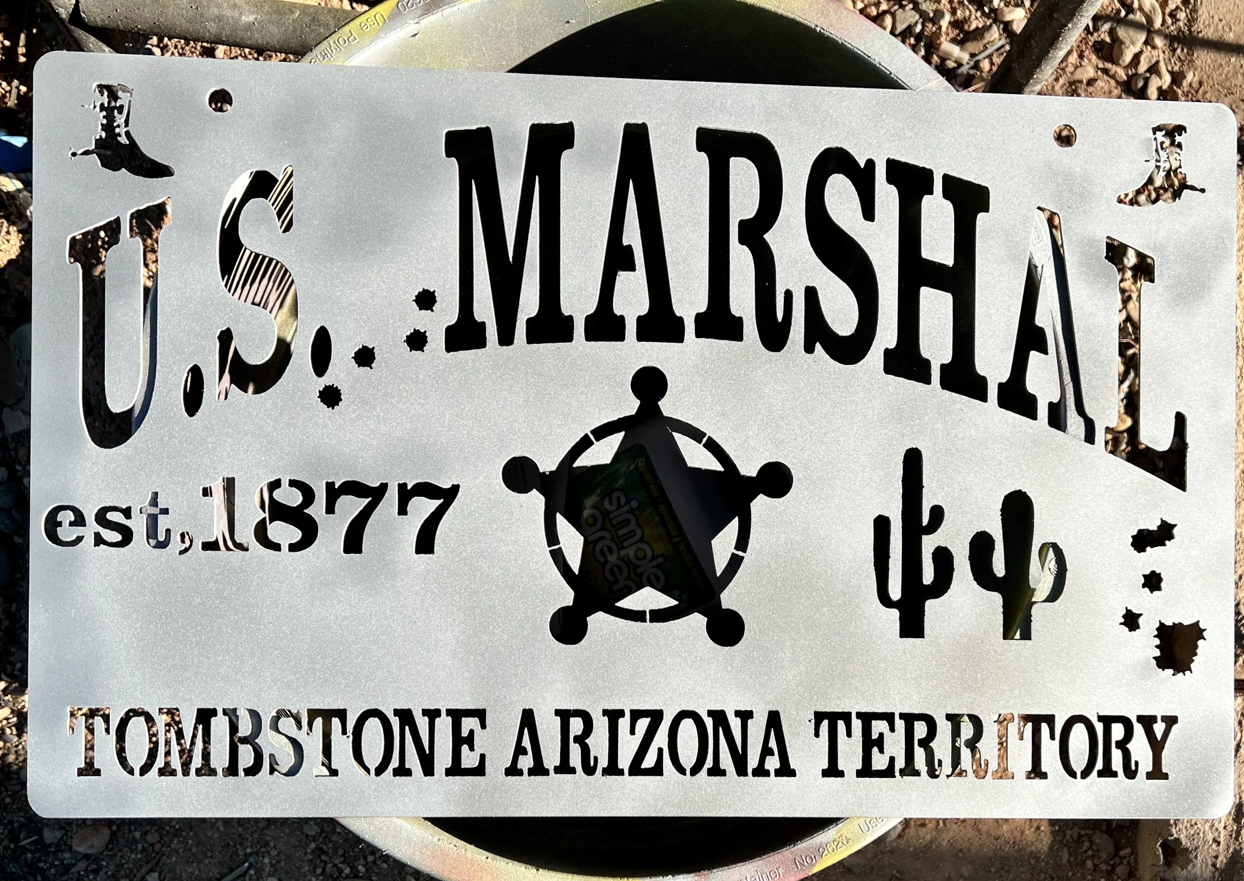 A metal sign with holes in the corners and cut-out text that reads 'TOMBSTONE ARIZONA TERRITORY' and the word 'MARSHA' at the top. The sign is placed on the ground among dirt and small rocks, with parts of a yellow hose and other outdoor equipment vi