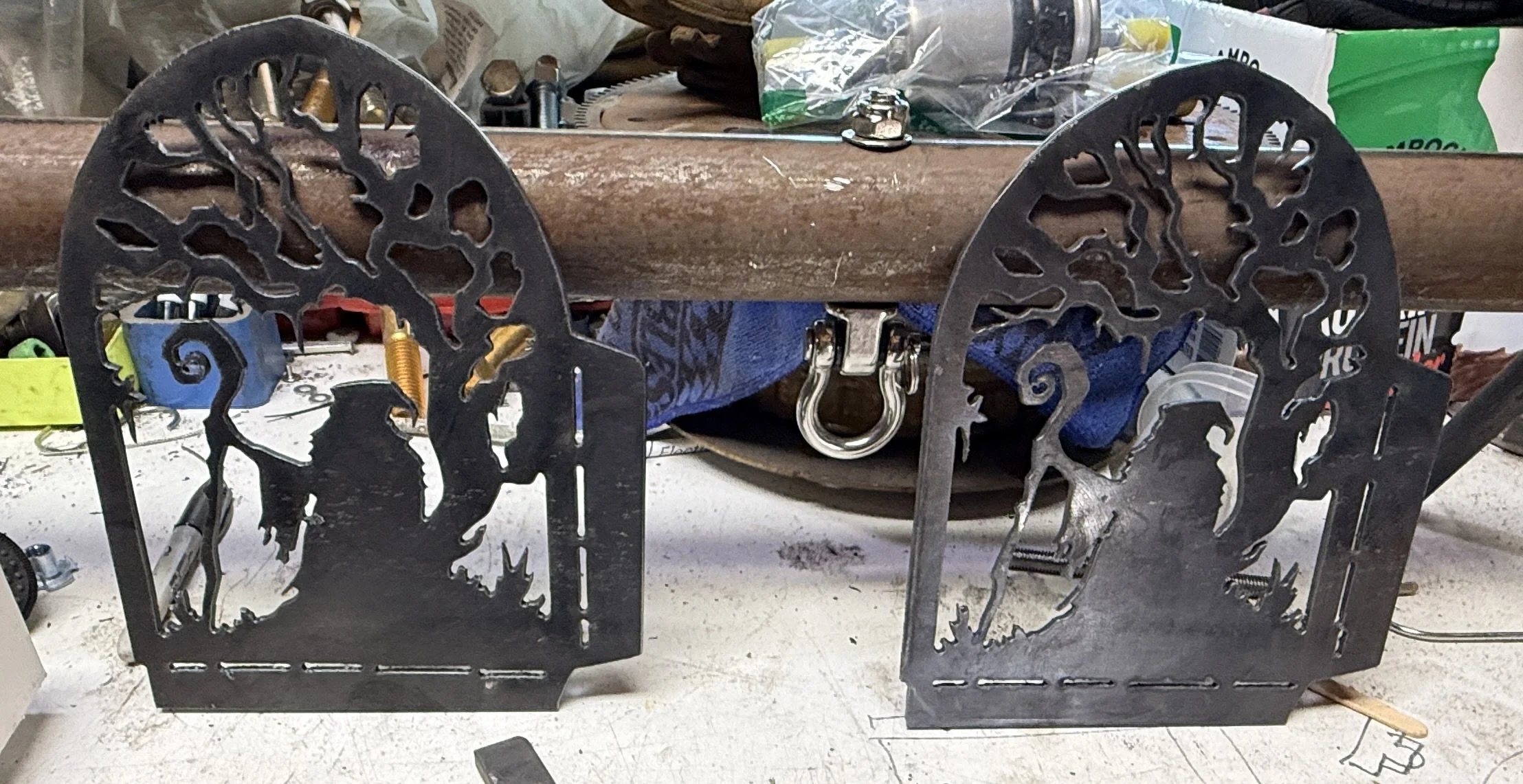 Metal cutouts with intricate designs of horses and trees on a workbench, with tools, hardware, and supplies in the background.