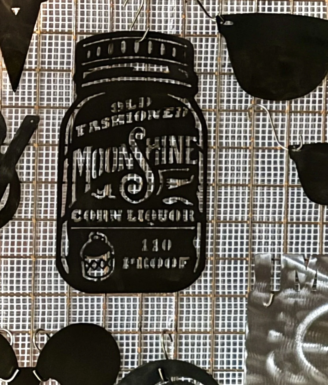 Decorative black metal sign shaped like a jar with text and a lantern illustration, surrounded by black objects, on a grid background.