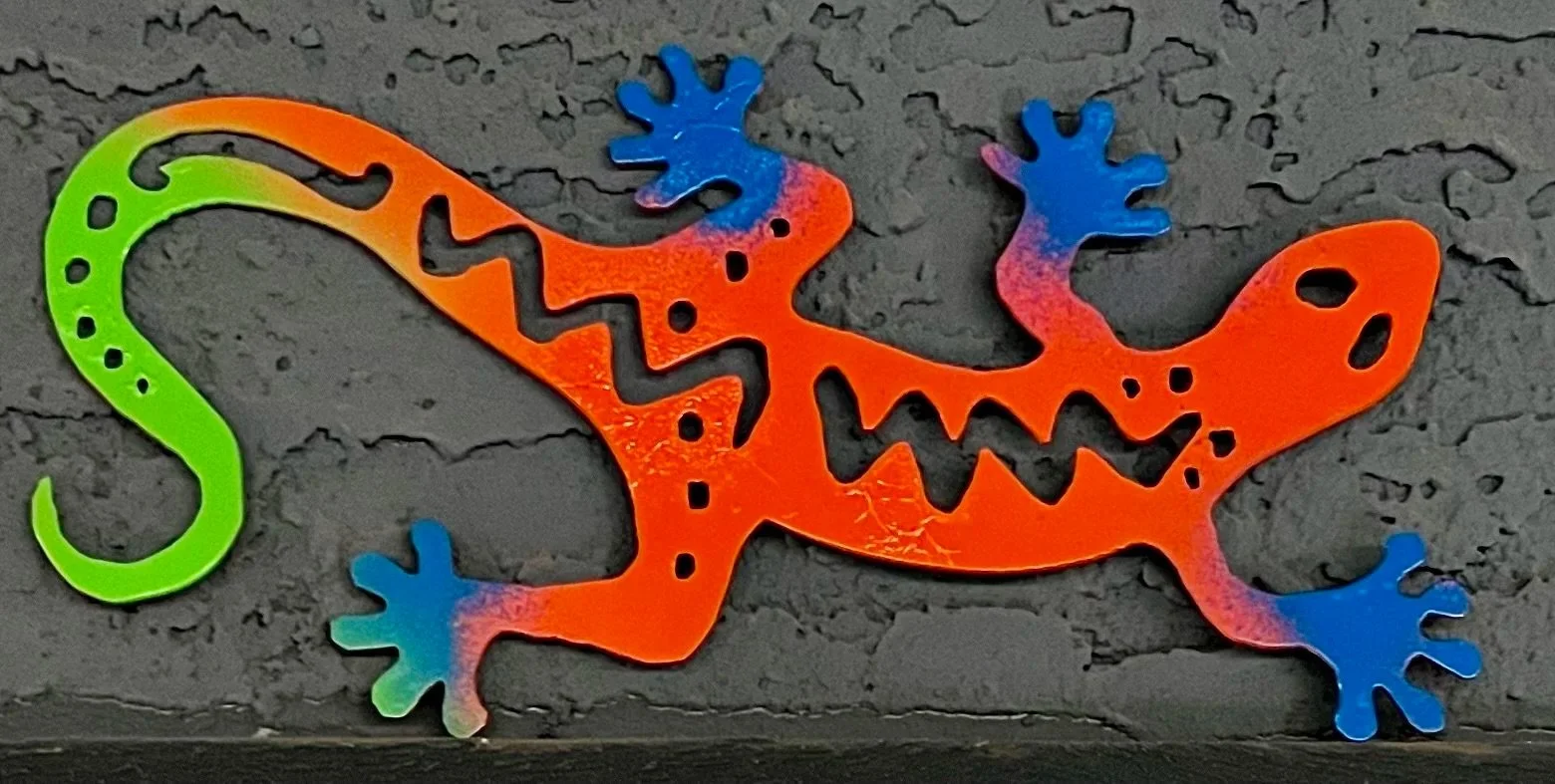 Colorful metal gecko decoration with bright orange body, blue and purple accents on the head and tail, and green tail, against a textured black background.