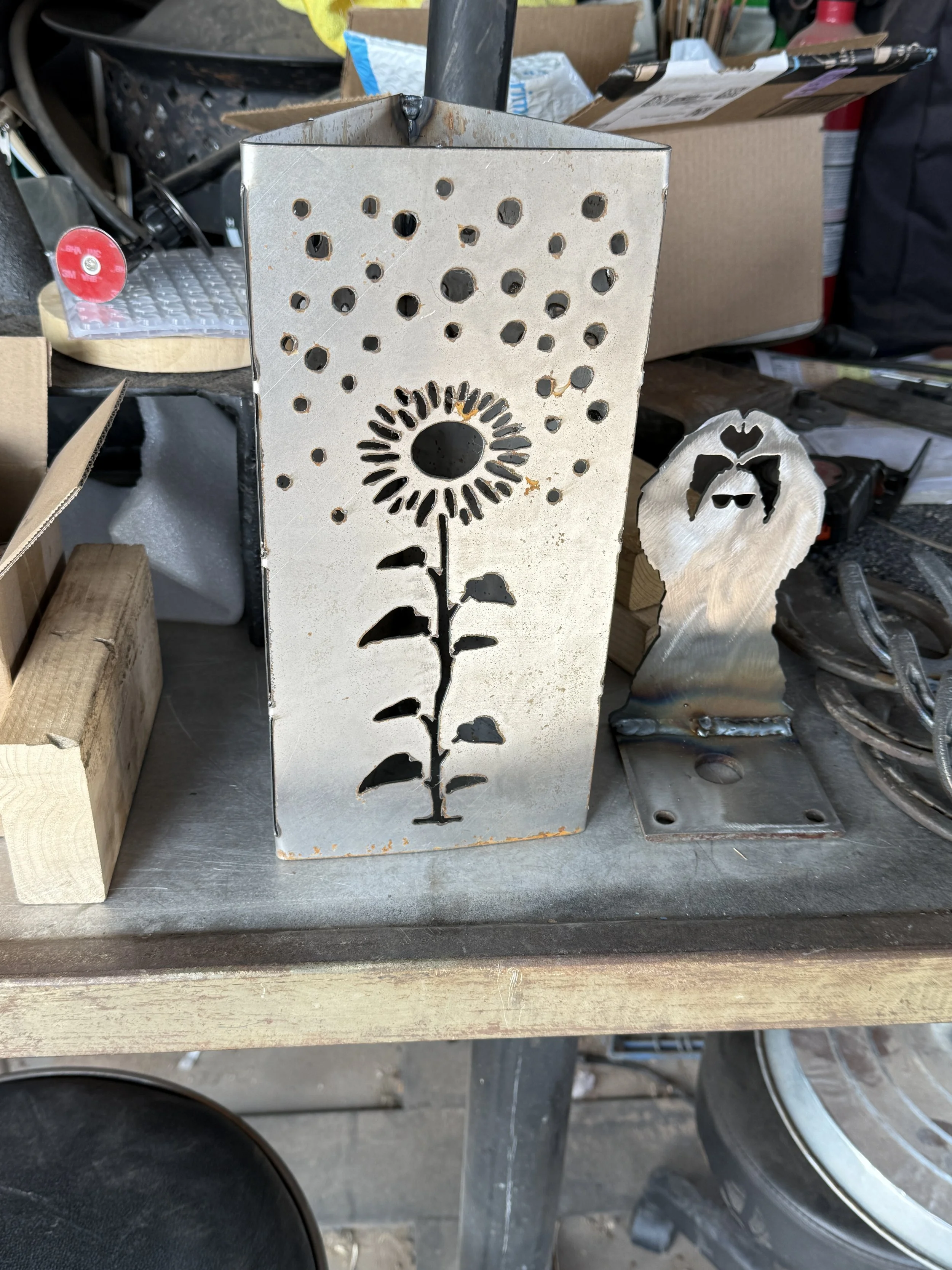 A metal piece with a sunflower design cutout, featuring a circular center and elongated petals, among holes of various sizes. It is placed on a workbench with other tools and materials in the background.