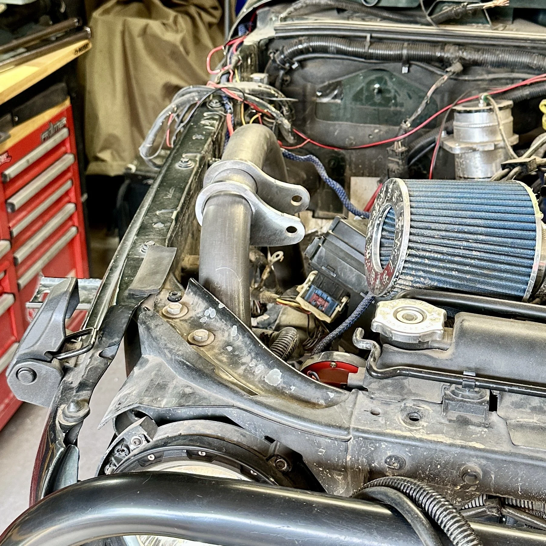 Car engine with exposed metal components, red and black wires, a large air filter, and various mechanical parts in a garage workshop.