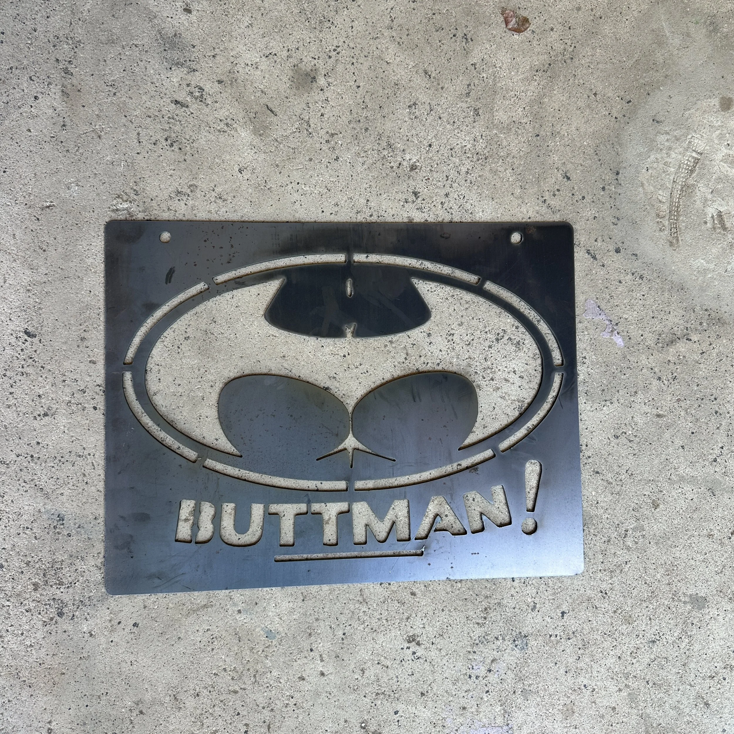 A metal grate with the word 'SUTTON' cut out, placed on a textured concrete surface, with part of a leaf visible at the top.