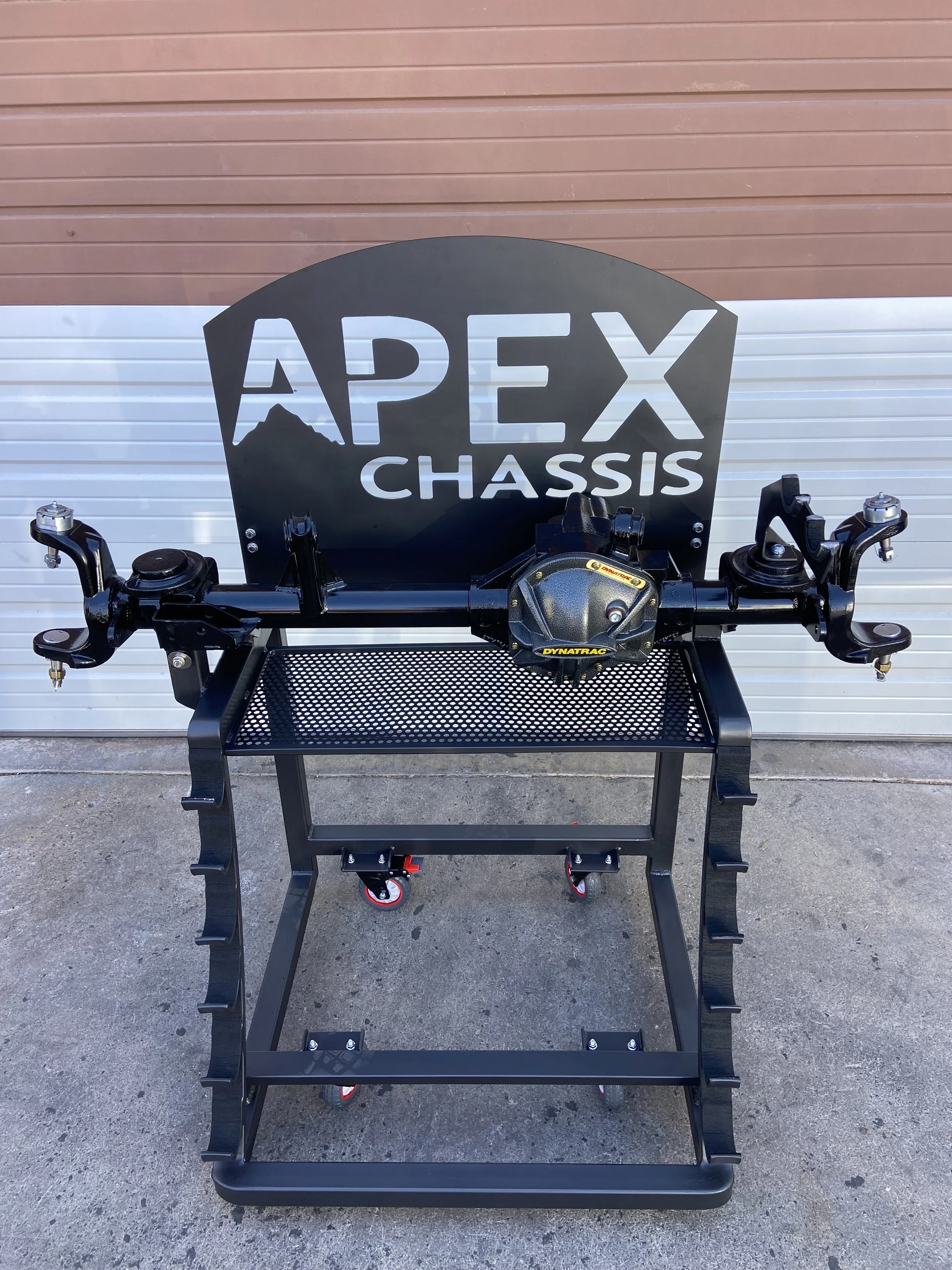 A race car steering wheel with a black headrest that says 'APEX CHASSIS' behind it, placed on a black metal stand against a white and brown door.