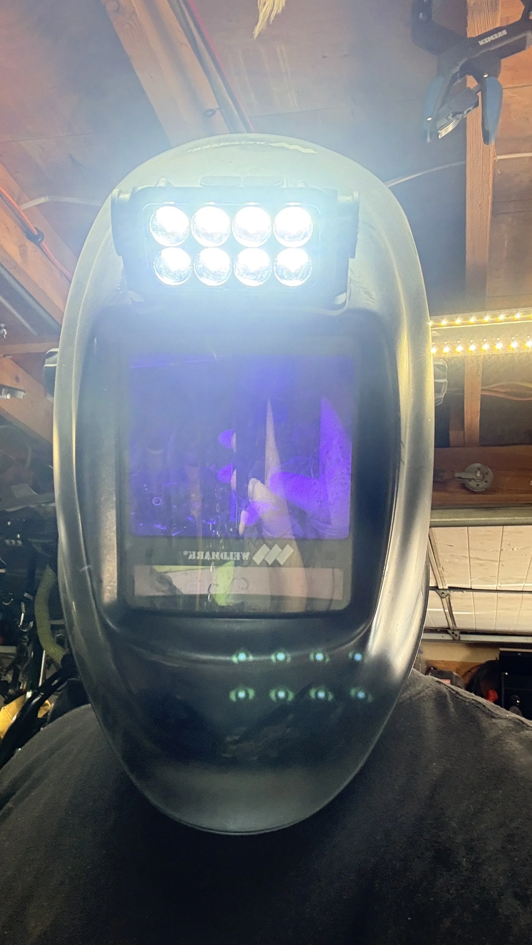 A handheld laser level with multiple bright LED lights, positioned on a person's lap inside a garage or workshop with tools and woodworking materials around.