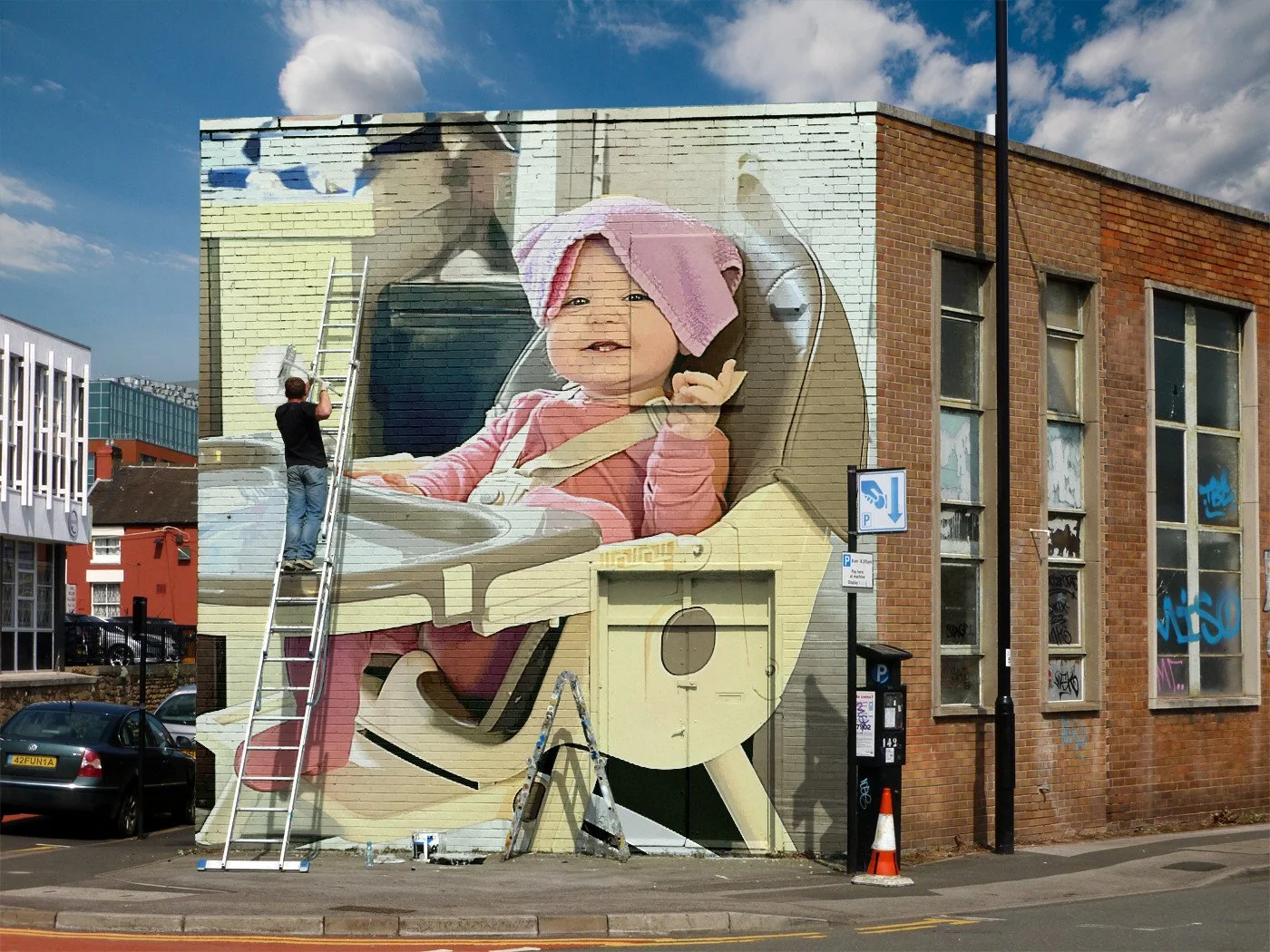 Mural of a baby in a car seat with a pink towel on its head, painted on the side of a brick building, with a person standing on a ladder working on it.