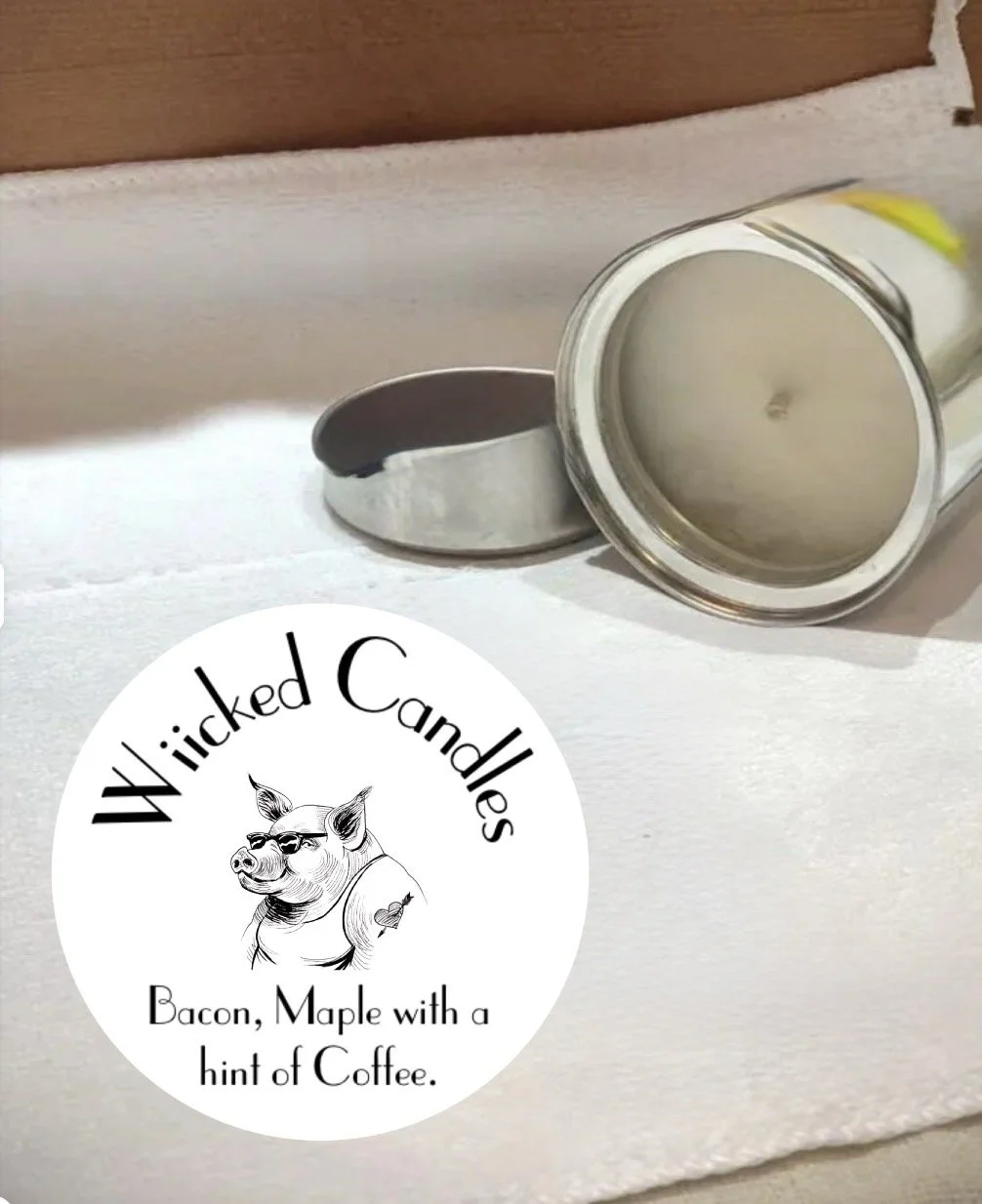 A round candle in a tin container with a lid next to it, placed on a white surface. There is a logo with a drawing of a pig wearing sunglasses and a tattoo, along with the text "Wicked Candles" and "Bacon, Maple with a hint of Coffee." 