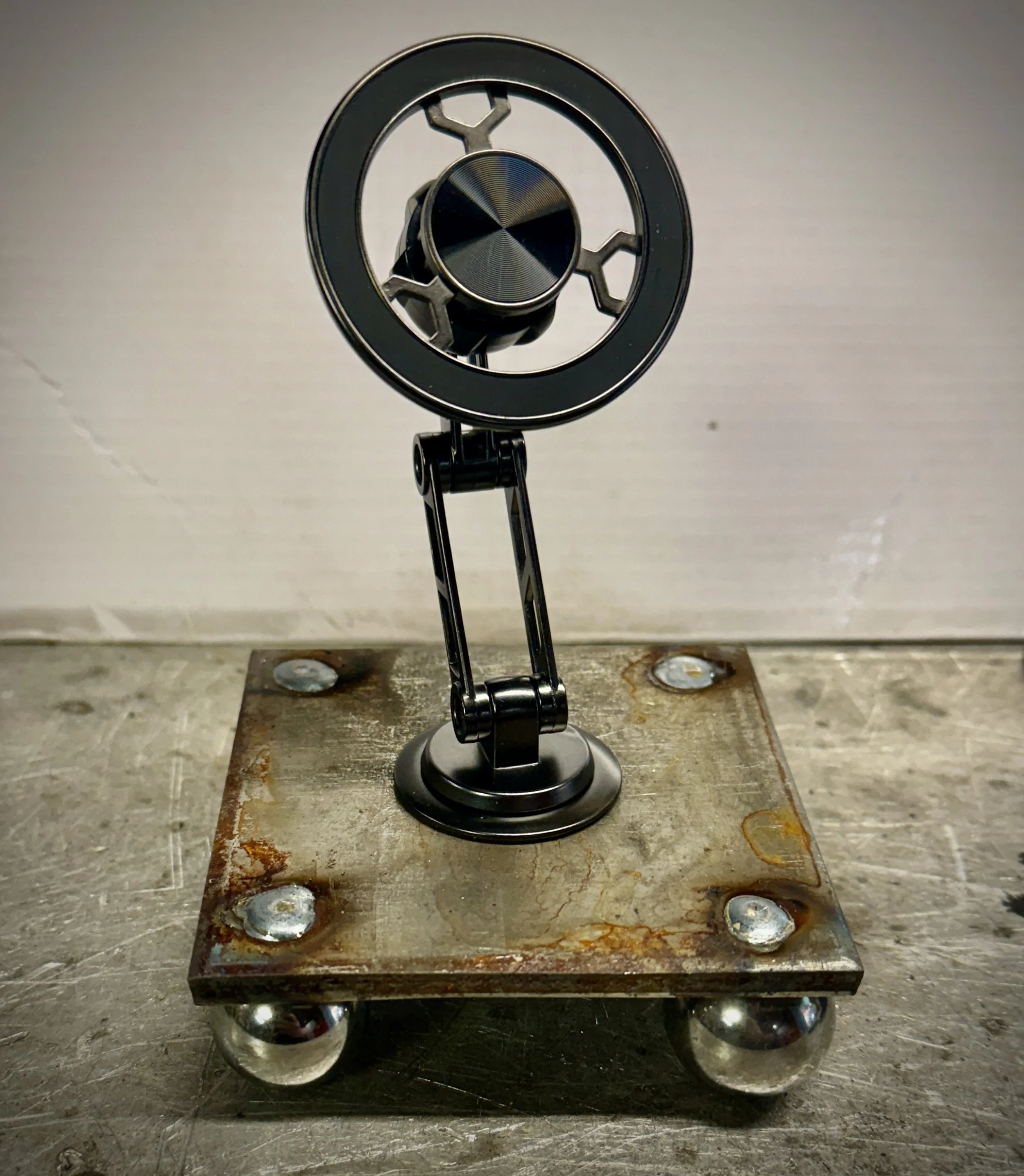 A close-up of a metal mechanical device with circular and hexagonal components, mounted on a rusted metal base with four spherical supports beneath.