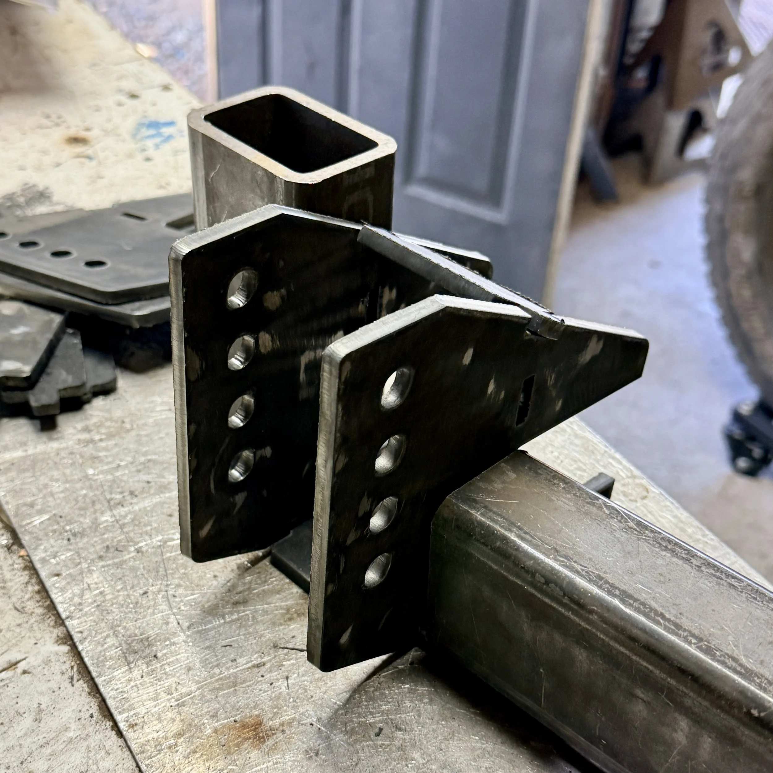 Close-up of metal bracket or support piece with holes, resting on a workbench or table in a workshop or metalworking environment.