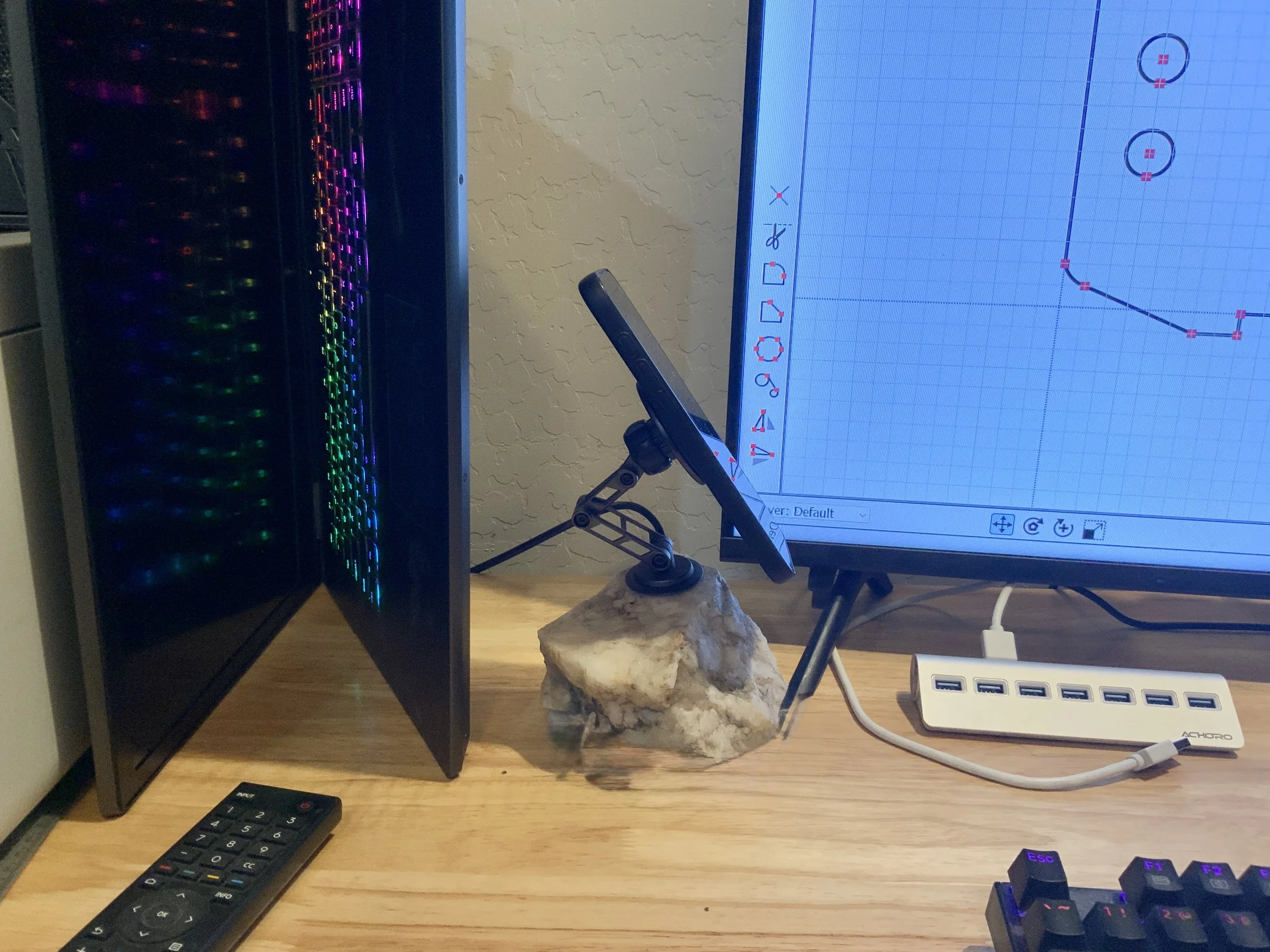 A computer desk setup shows a gaming keyboard, a calculator, and a monitor displaying a graph. To the left, there's a desktop computer case with colorful RGB lighting. A stand holds a smartphone in front of the monitor, which is placed on a stone-lik