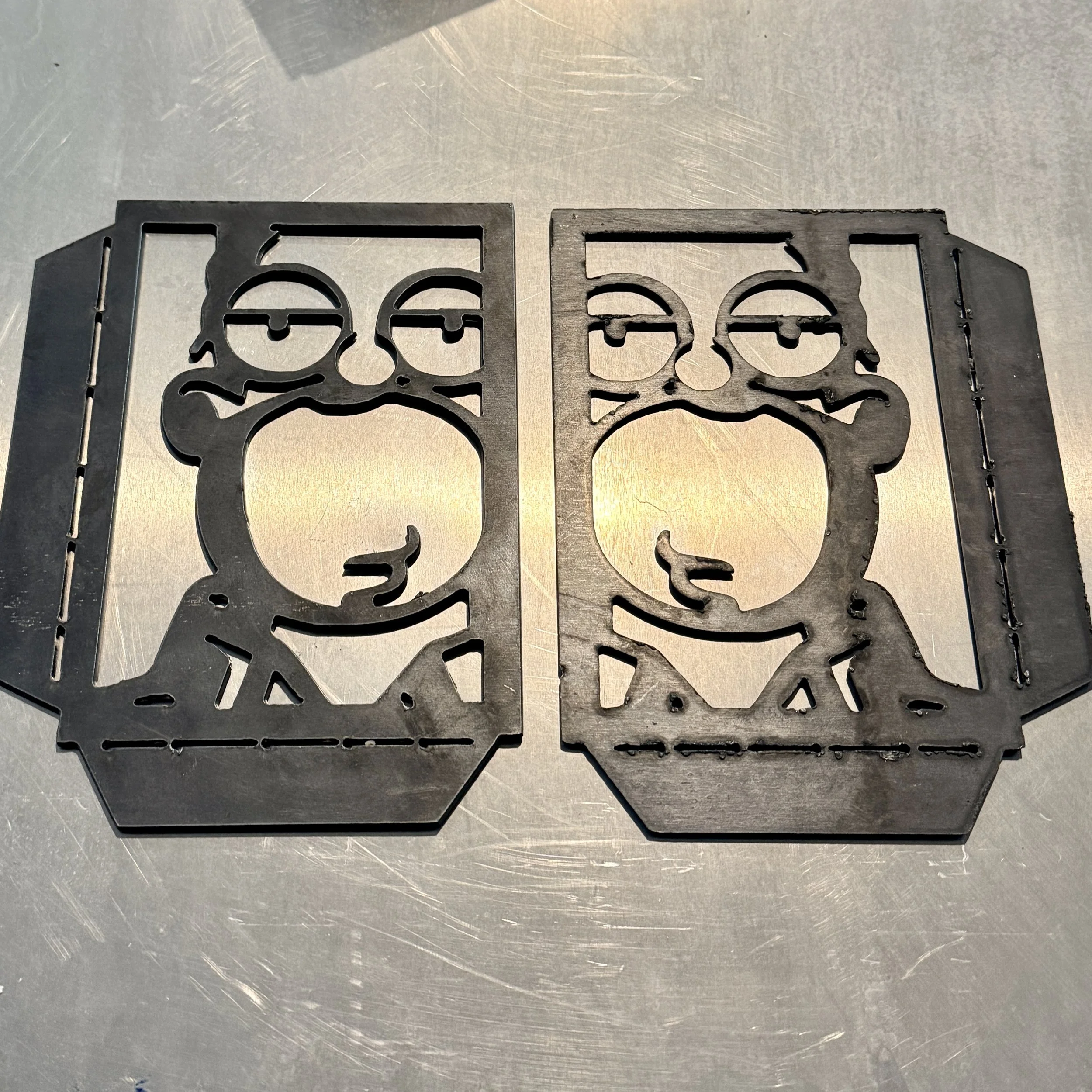 Two metal laser-cut sheets with a skull and crossbones design, placed on a metallic surface.