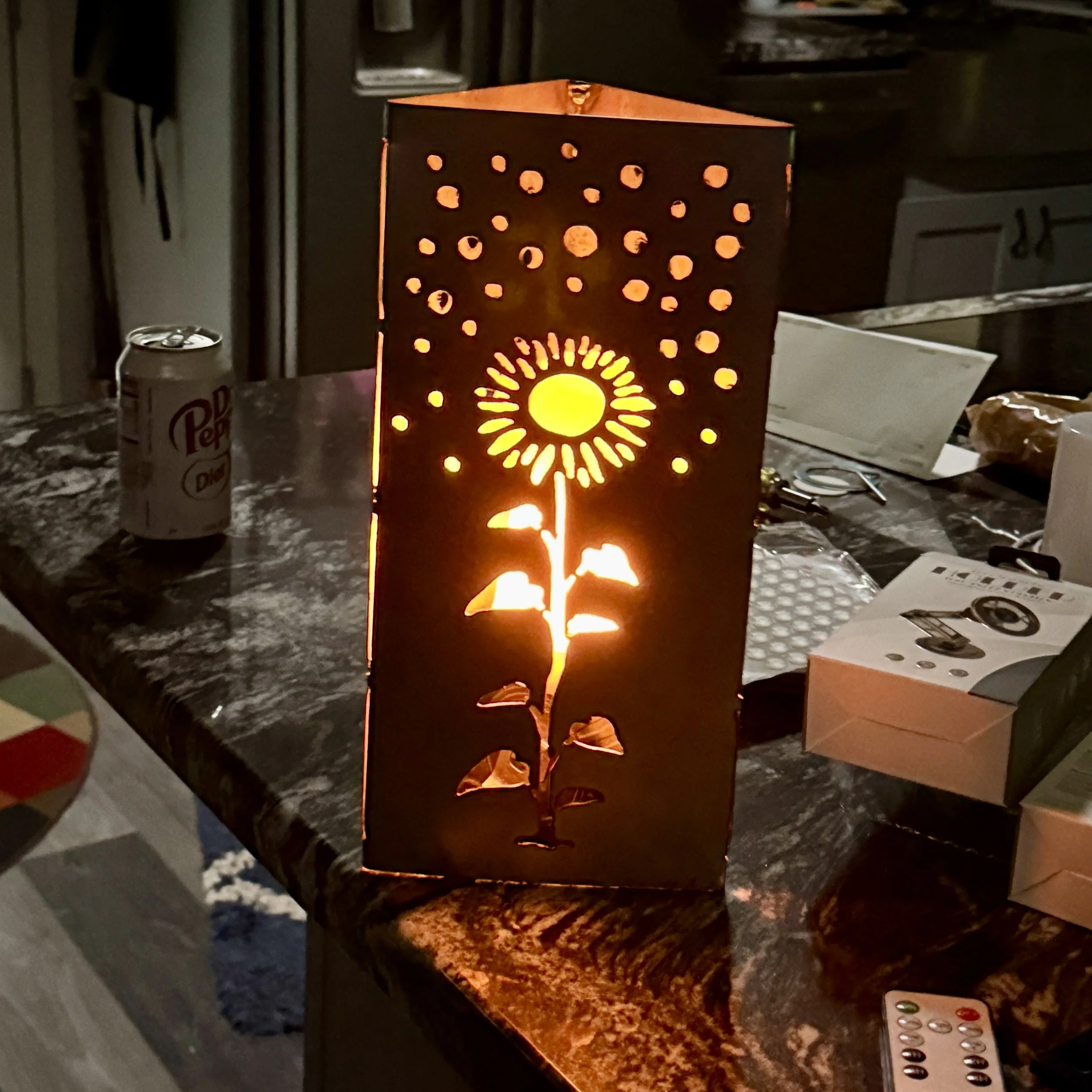 A lit decorative candle holder with circular cutouts resembling a sunburst pattern, placed on a granite countertop in a dimly lit room with various objects nearby.