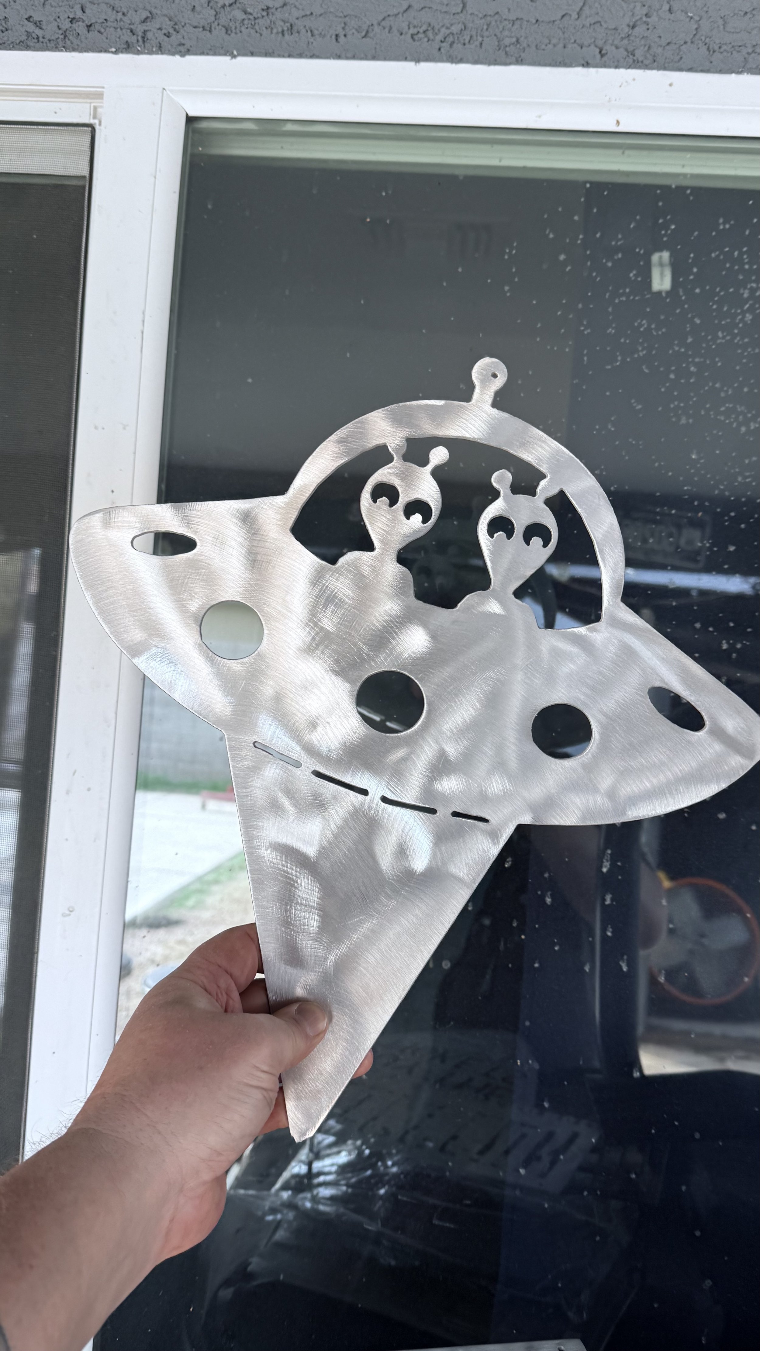 A person holding a metal decorative piece shaped like a butterfly with two alien heads inside it, outlined with holes and cutouts, in front of a window.