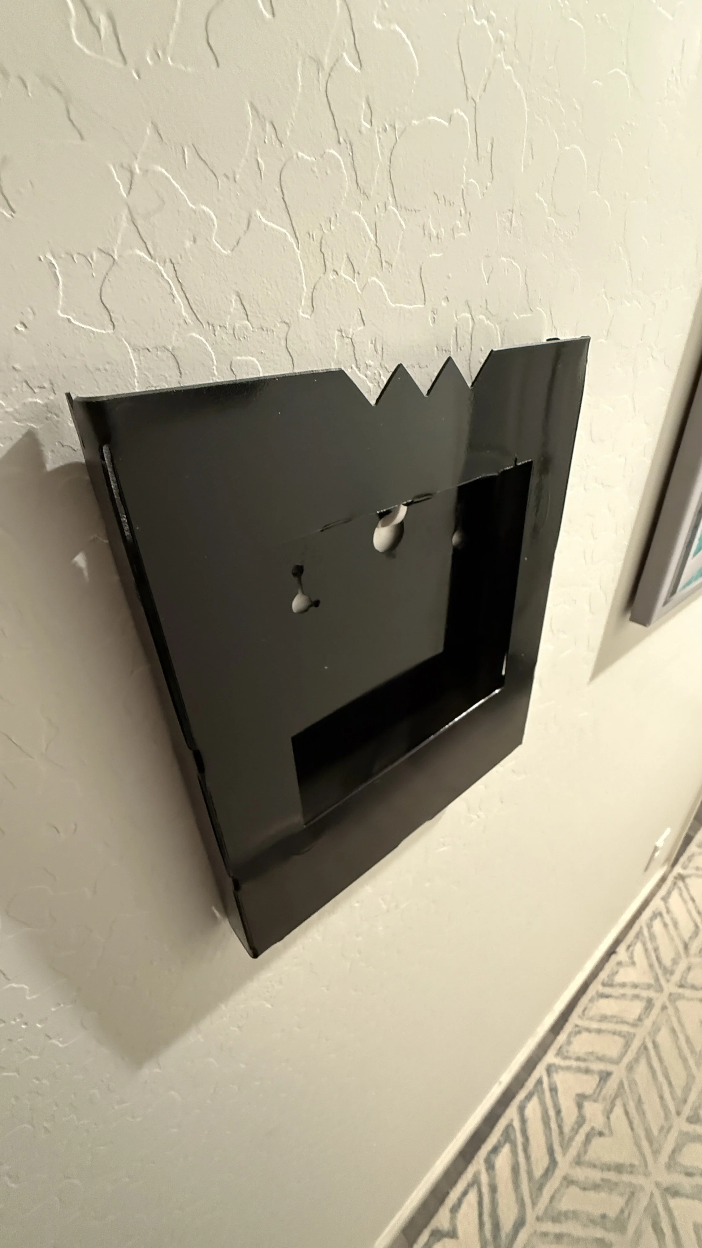 Black wall-mounted cabinet with an open front, empty inside, attached to a textured off-white wall, beside a patterned tile floor.