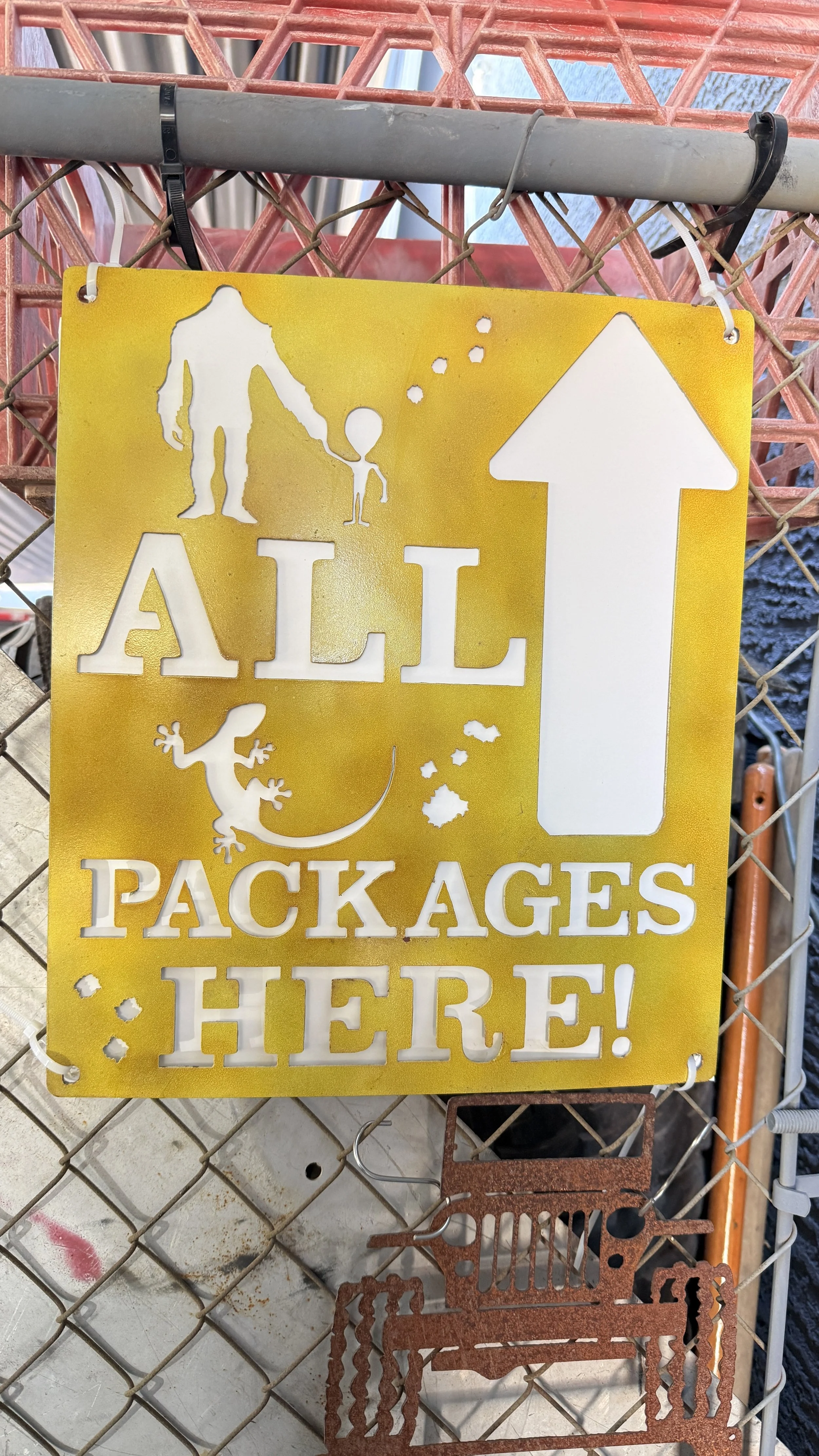 Sign with cutouts of a man and child holding hands, a gecko, an arrow pointing up, and the words 'ALL PACKAGES HERE!'. The sign is hanging on a chain-link fence.