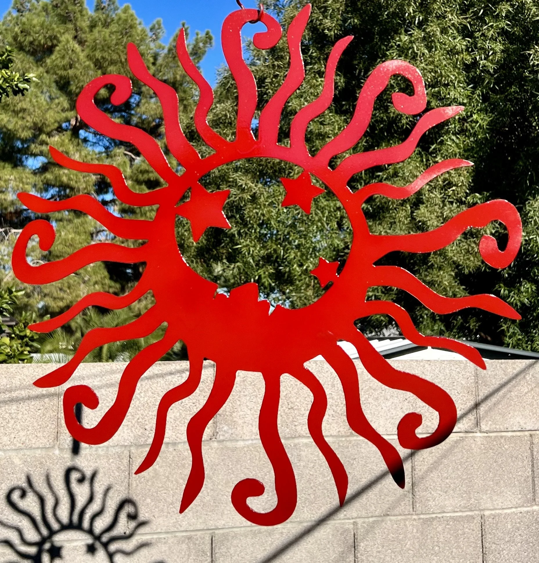 Red metal sculpture shaped like a stylized sun with swirling rays and stars, mounted on a stone wall, with trees and blue sky in the background.