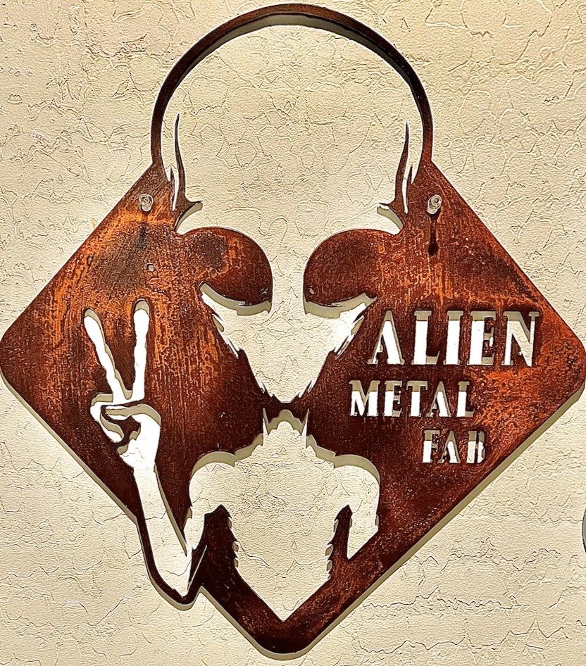 Wall art of a space-themed metal_sign with an alien holding a peace sign, accompanied by the words 'Alien Metal Fab'.