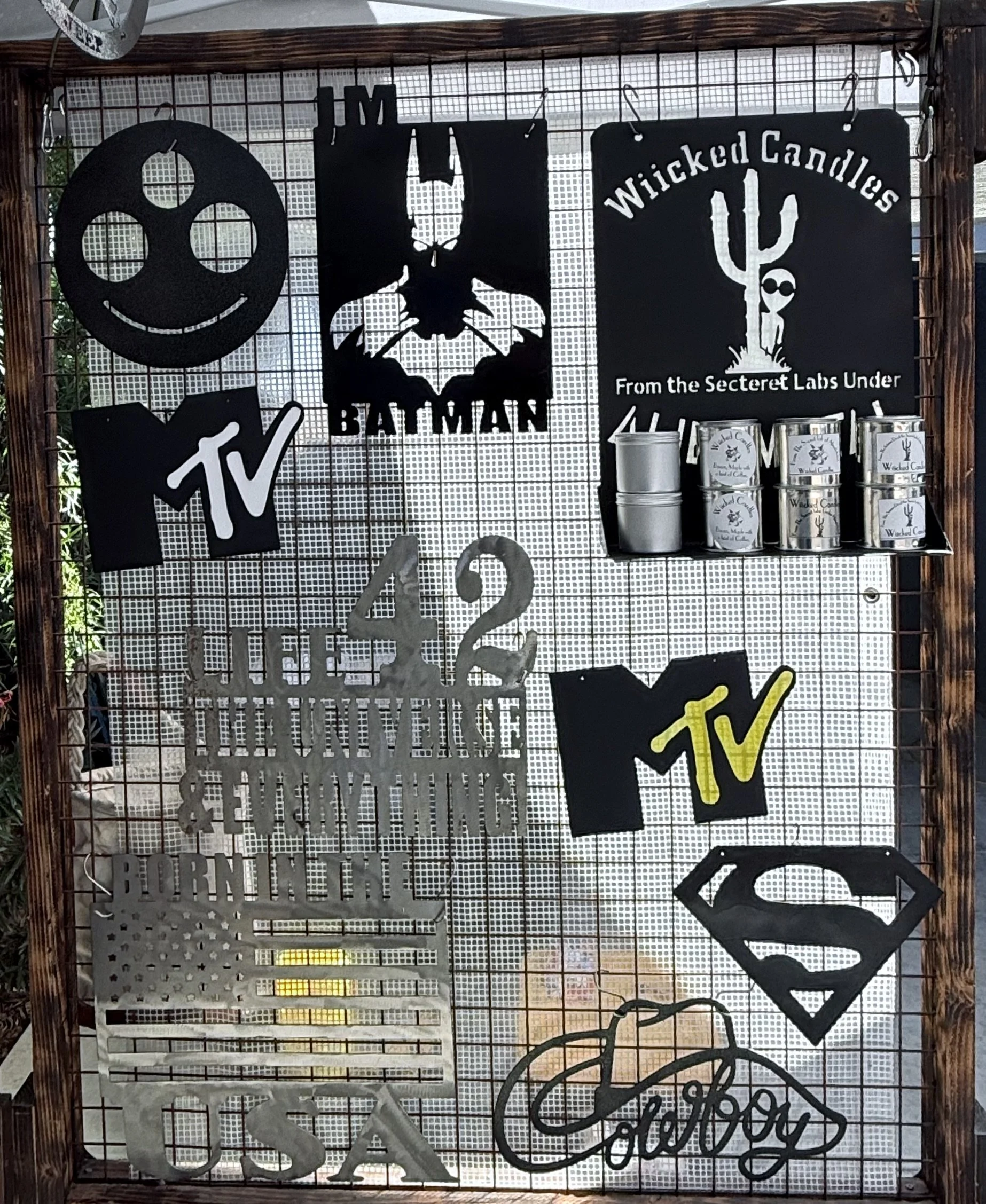 Various black metal cut-out signs and decorations hanging on a wire mesh grid display. The signs include the MTV logo, a smiling face with three circles for eyes, a Batman logo, a 'Wicked Candles' sign with a cactus and skull design, and other decora