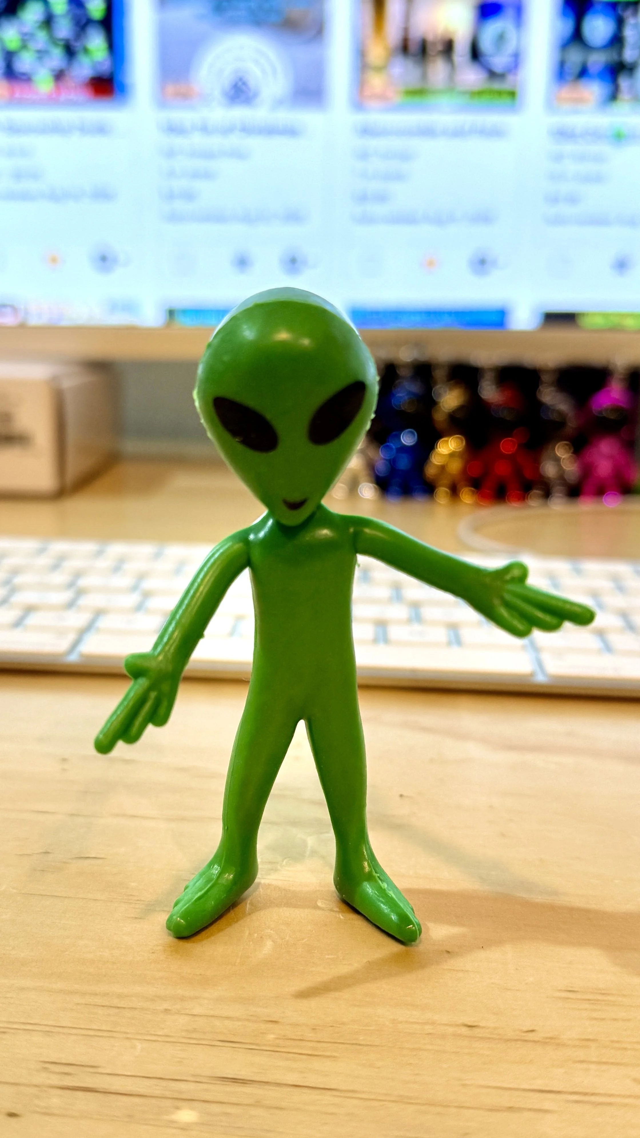 A small green alien figurine with large black eyes and a long neck, standing on a wooden surface with a blurred computer monitor and colorful objects in the background.