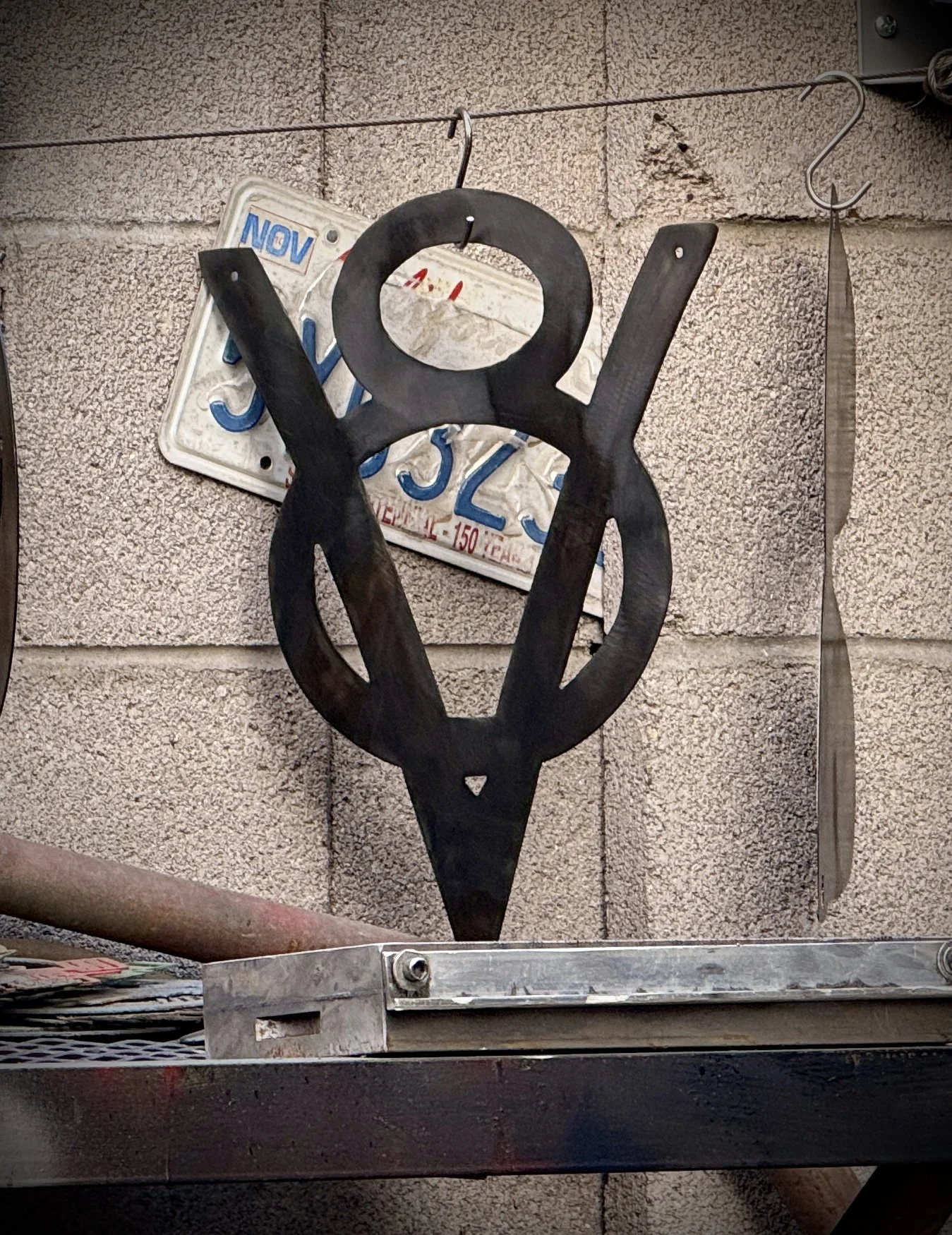A metal art piece resembling the Greek letter Omega, mounted on a stone wall. Behind it, a plastic license plate with the characters 'S 2 3' is visible, slightly obscured. To the right, a thin, flexible metallic strip is attached to the wall.