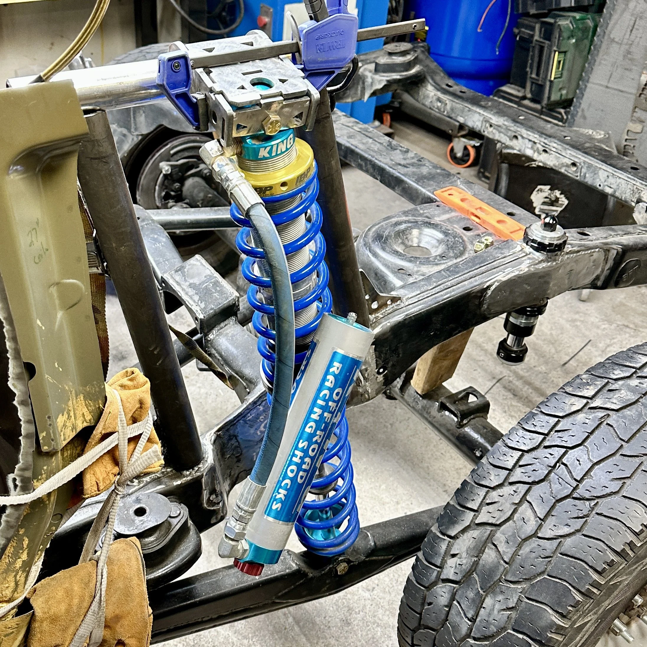 An automotive chassis with a suspension system that includes a blue coil spring and shock absorber. The chassis appears to be in the process of assembly or modification in a workshop, with a tire, a towel, and various tools nearby.
