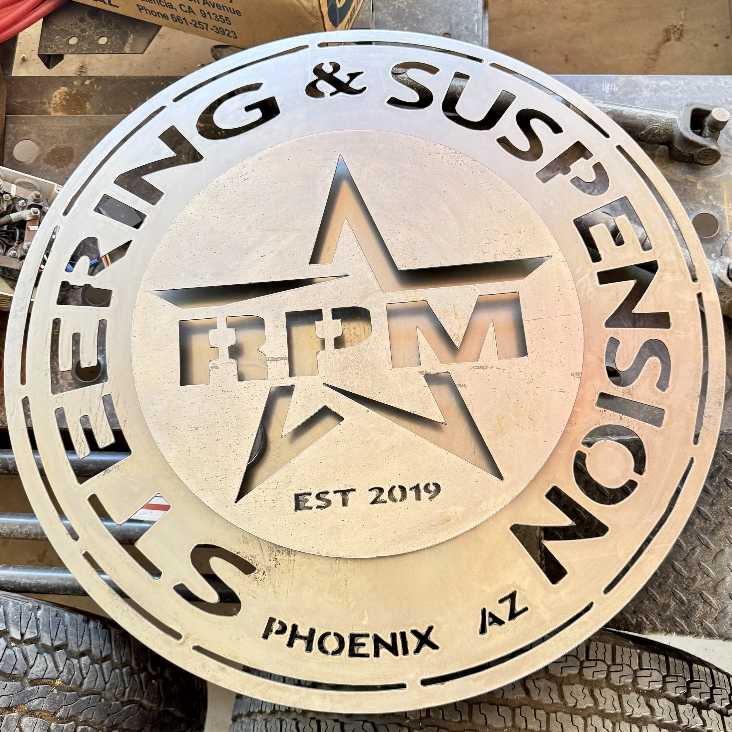 A circular metal sign with the words 'LUX & SUNSET PHOENIX' and 'EST 2019' engraved around a central star logo, placed on a worktable with tools and a cardboard box nearby.