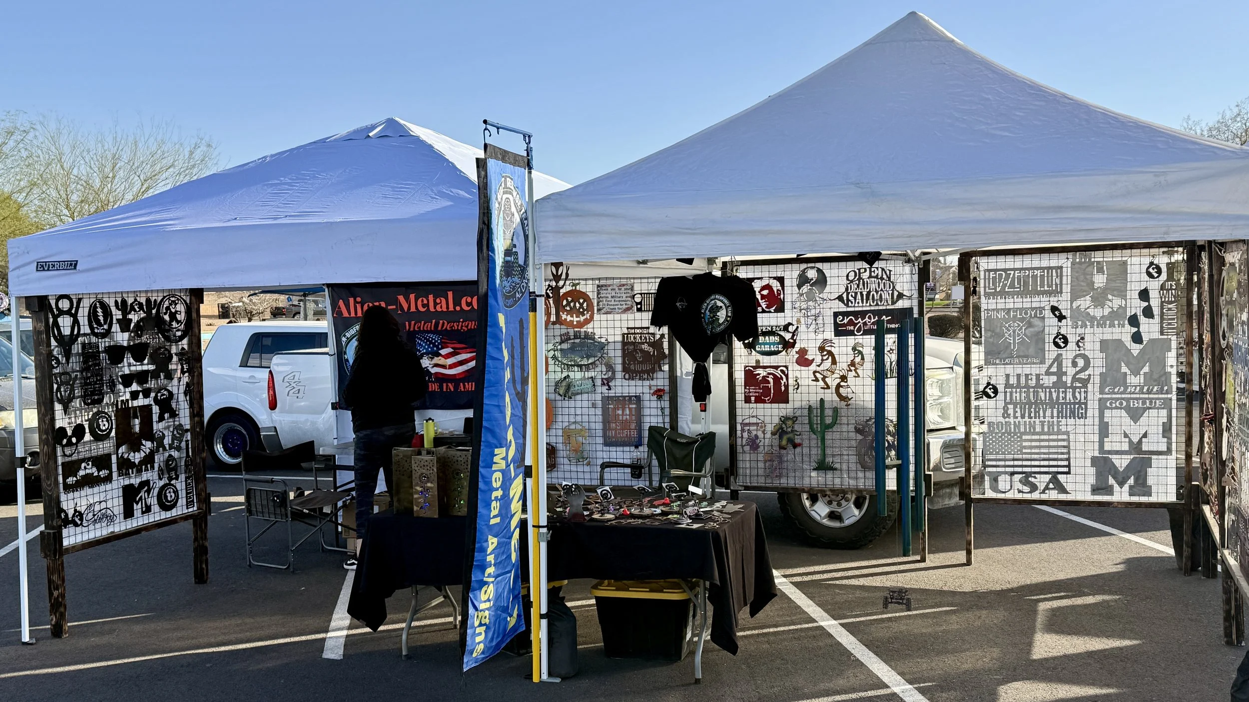 Outdoor market stall with various metal and wood crafts, including decorative signs and cutouts, housed under a white canopy in a parking lot.