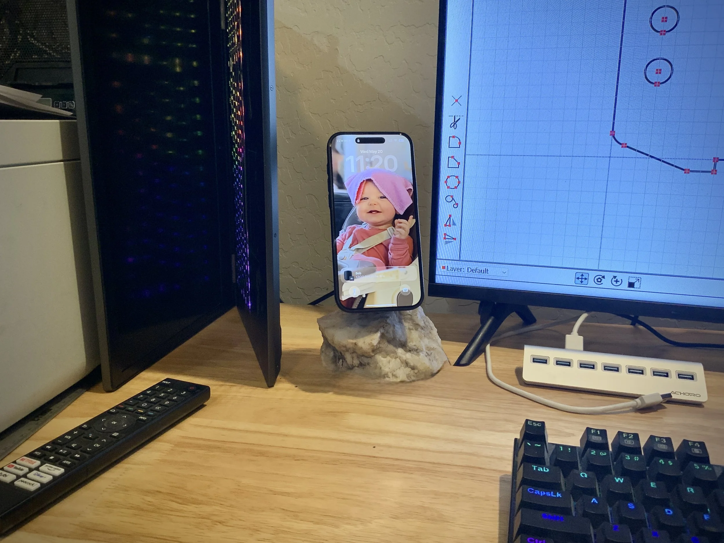 Computer desk with a monitor displaying a hockey game, a keyboard, a mouse, a smartphone showing a baby with a pink towel on its head, a computer tower with rainbow LED lights, and a remote control.