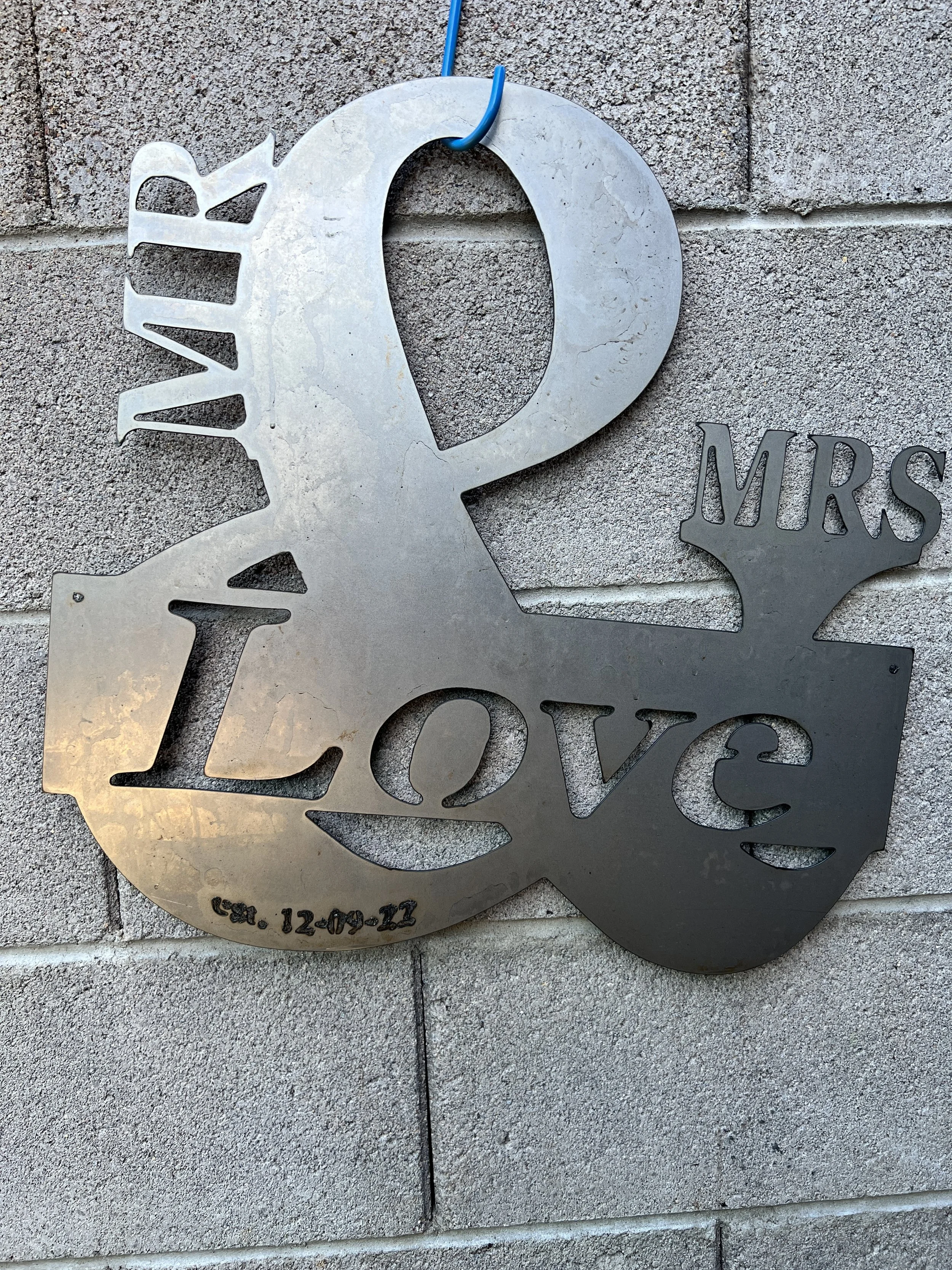 Metal wall art featuring the words 'LOVE' and 'MRS' with a heart and date 'est. 12-09-11' against a textured brick wall.
