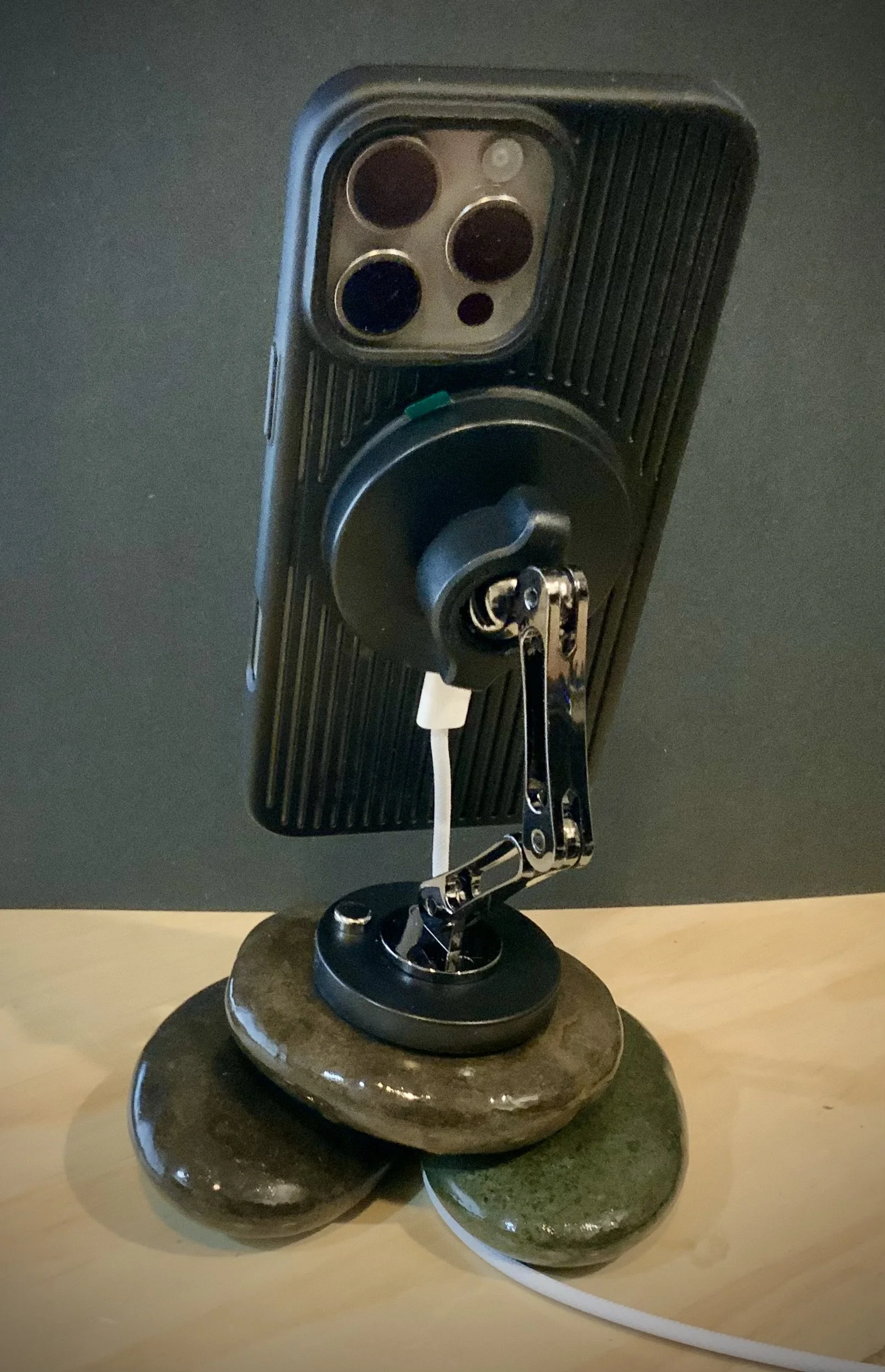 Close-up of a smartphone mounted on a flexible stand, positioned behind three smooth, polished stones of varying sizes and colors on a wooden surface.