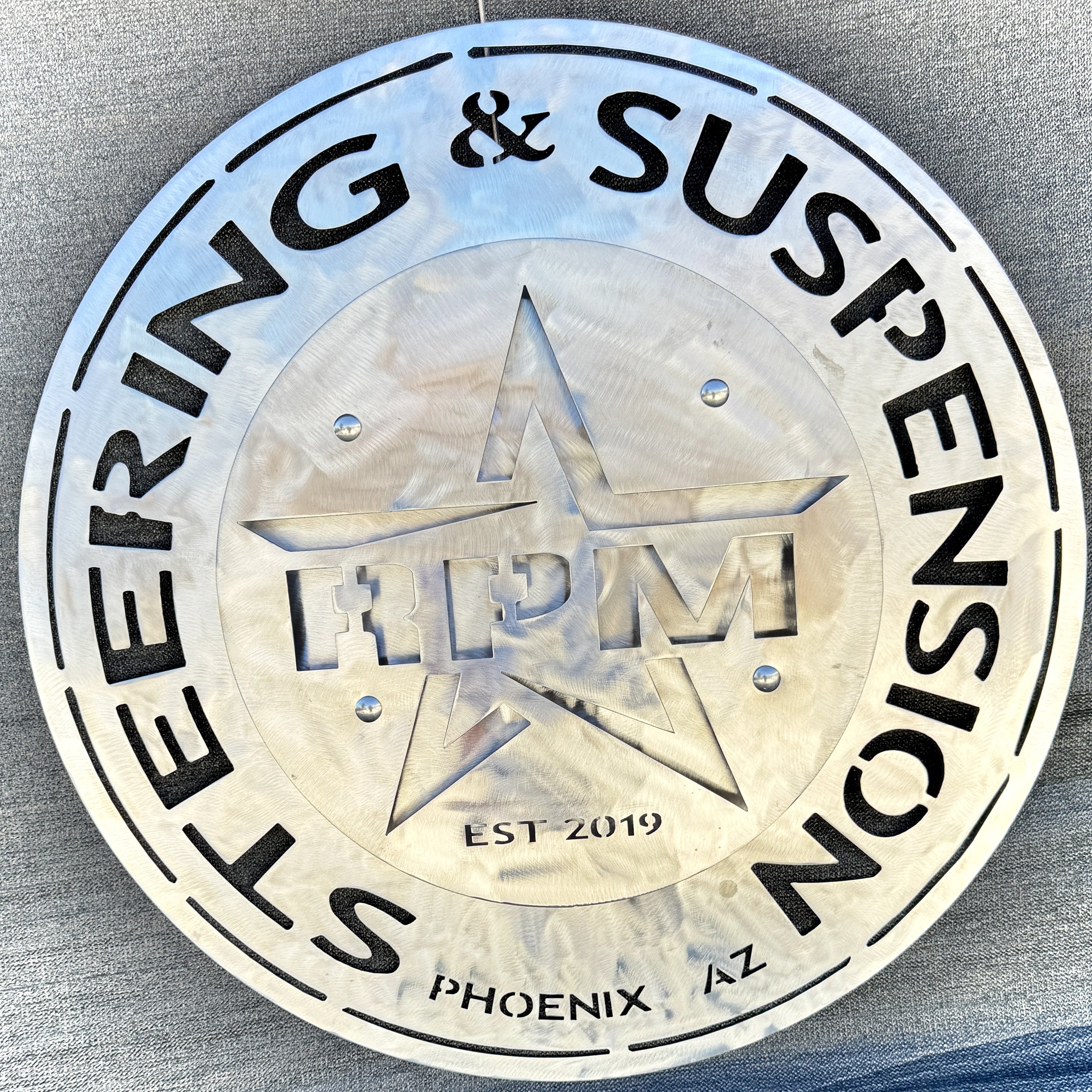 Silver circular sign with black cut-out text that reads 'Phoenix, AZ' at the bottom and 'EST 2019' below. The background is a gray textured surface.