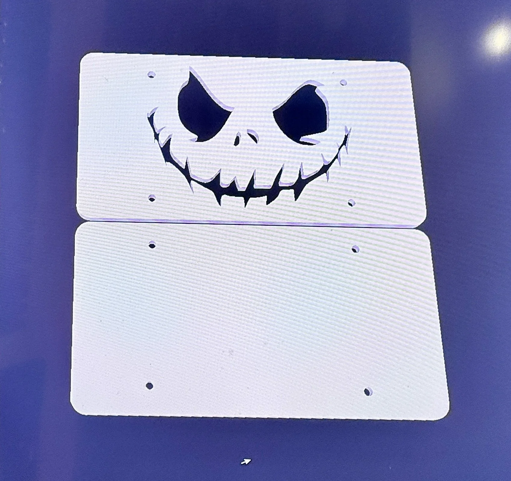 A metal license plate with a cutout design of a grinning skull face, mounted on a dark blue surface.