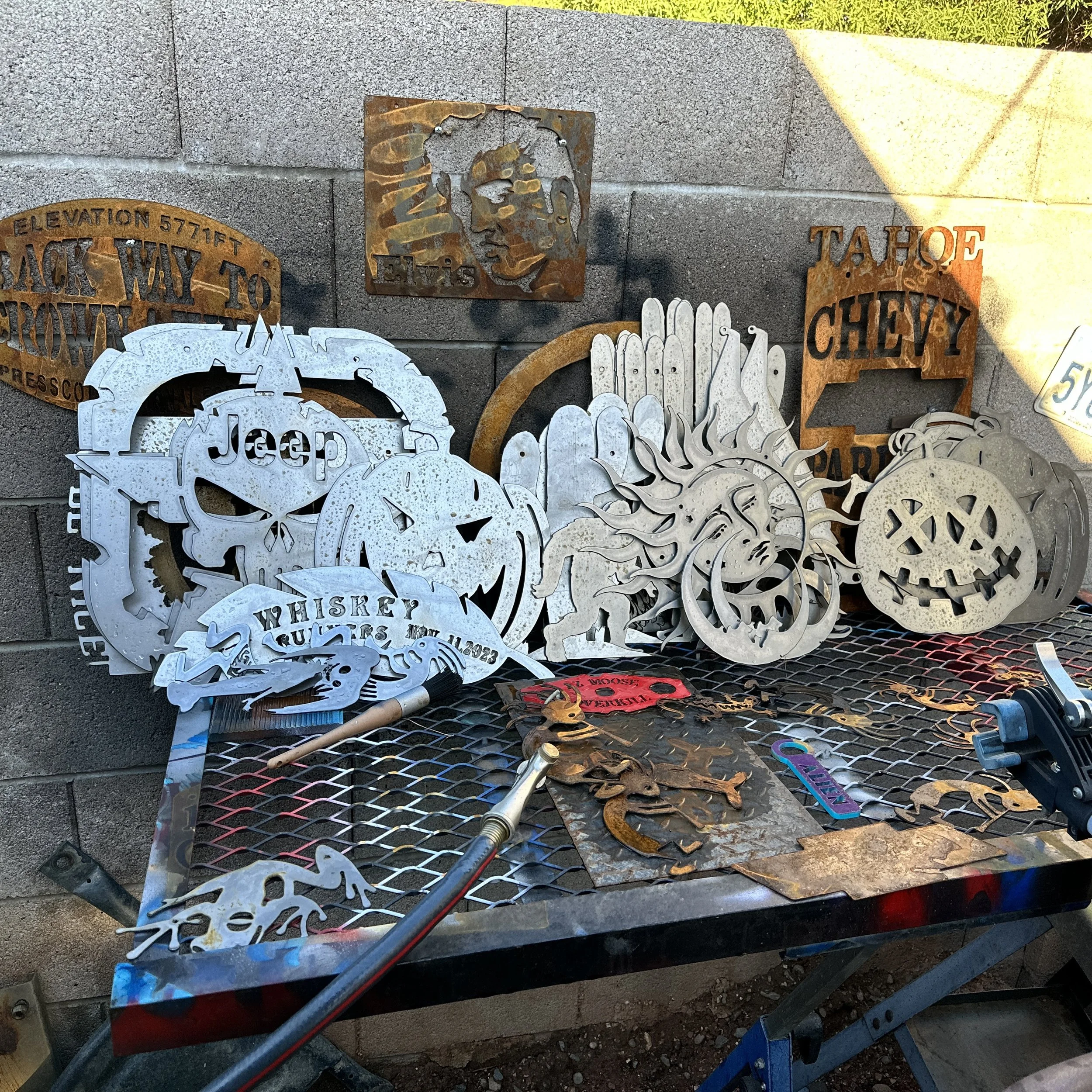 Collection of metal art pieces with various designs, including a face profile, a skull, and words like 'Jeep' and 'Tahoe,' arranged on a metal table outdoors.