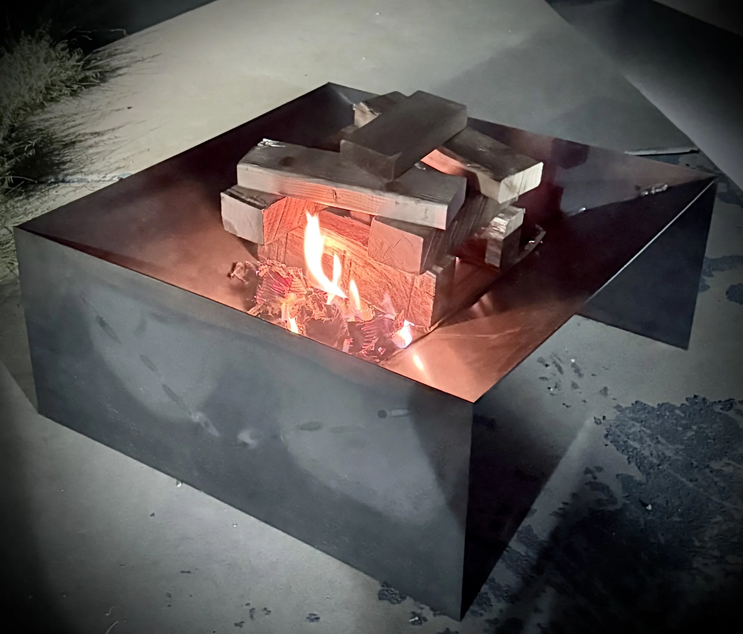 A metal fire pit with several pieces of chopped wood burning inside, with a small flame visible and some smoke rising. The fire pit is on a concrete surface, and there is some grass and plants in the background to the left.