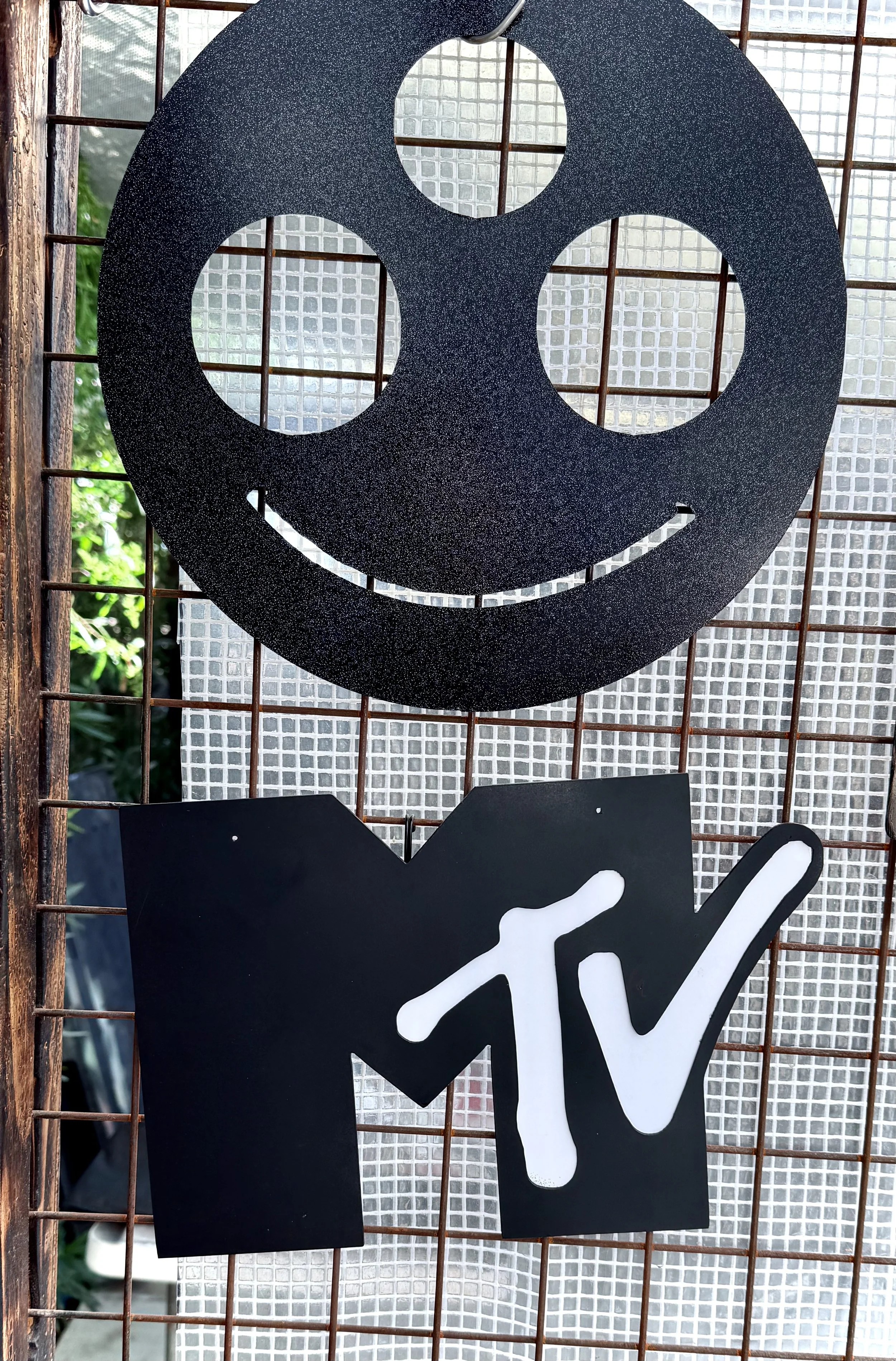 Close-up of a black smiley face sign with a rounded face and two circular eyes, attached to a rust-colored wire grid with a glass block wall and some greenery in the background.