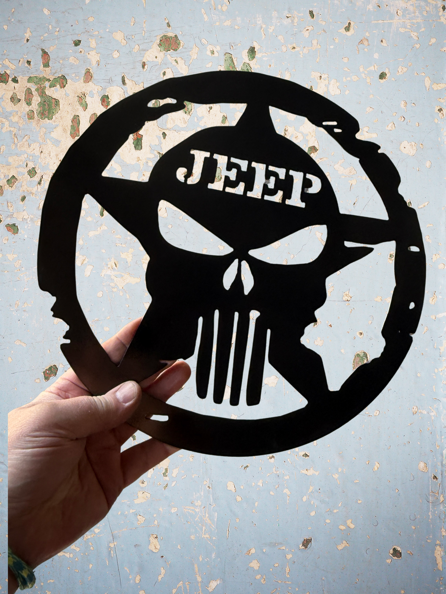 A black metal cutout featuring a skull with a helmet and the word 'JEFF' at the top. The person holding it is wearing a multicolored braided bracelet on their wrist.