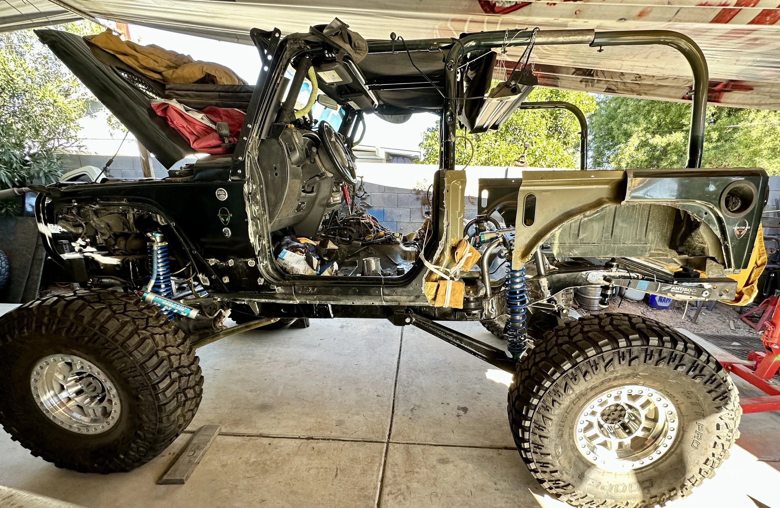 An off-road vehicle with large tires and a partially disassembled body in a garage, with some parts and tools visible, and trees seen outside.