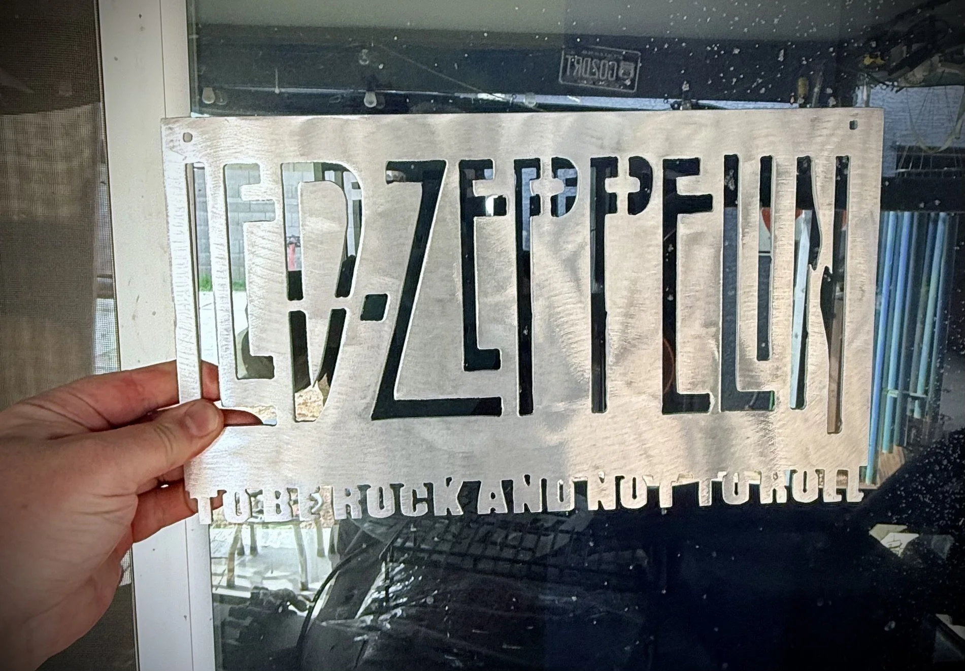 A person's hand holding a metal stencil with cut-out letters and numbers reading "LED 7ZFHISPEN" in front of a glass door, with some objects visible through the glass.