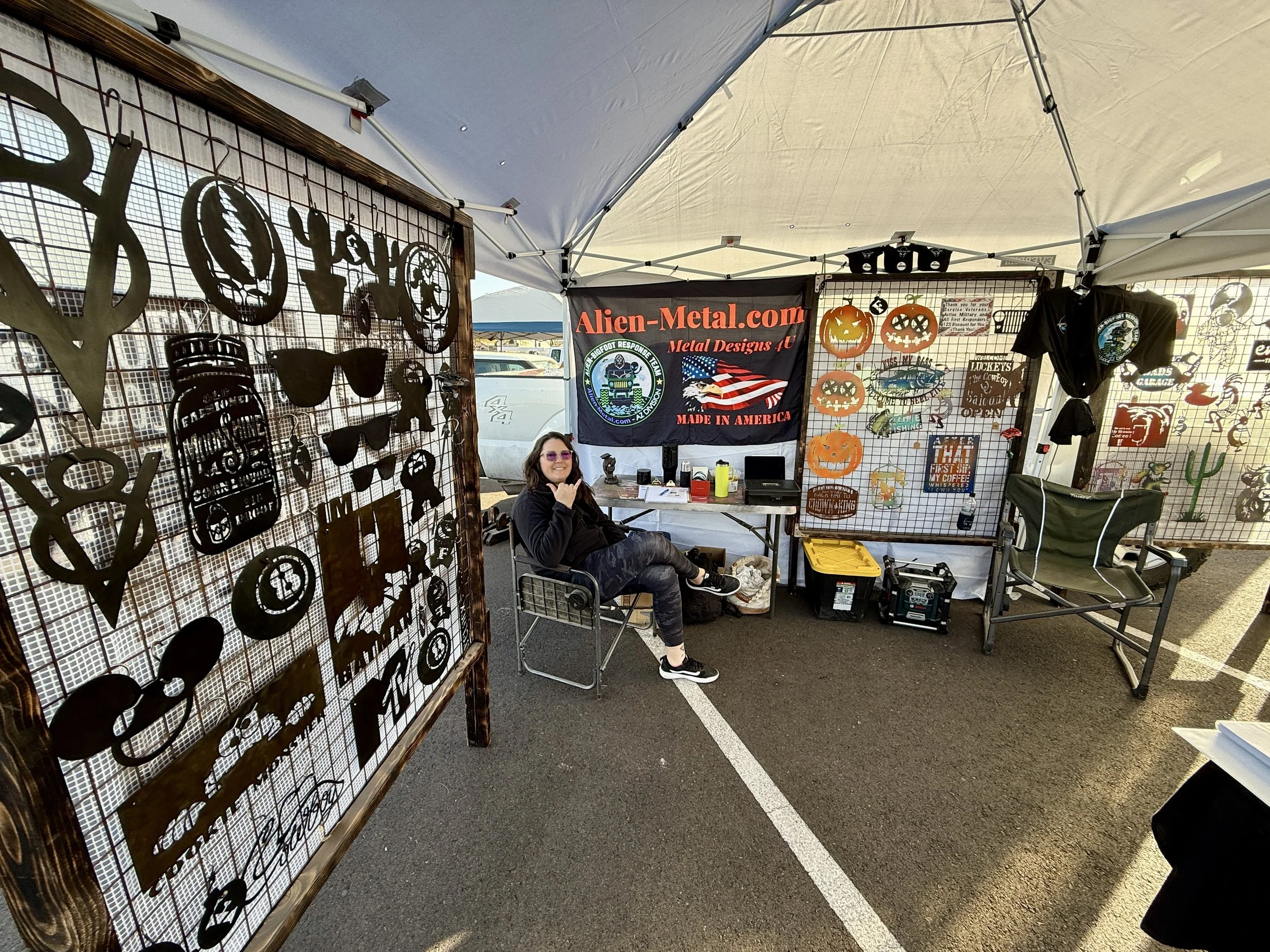 At an outdoor market stall, a woman with glasses and dark hair sits and smiles, surrounded by various metal and Halloween-themed decorations on display, including pumpkins and skeletons, under a white canopy.